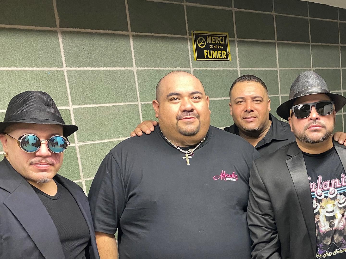 Four men standing together against a green tiled wall, some wearing sunglasses and hats, with a sign above them.