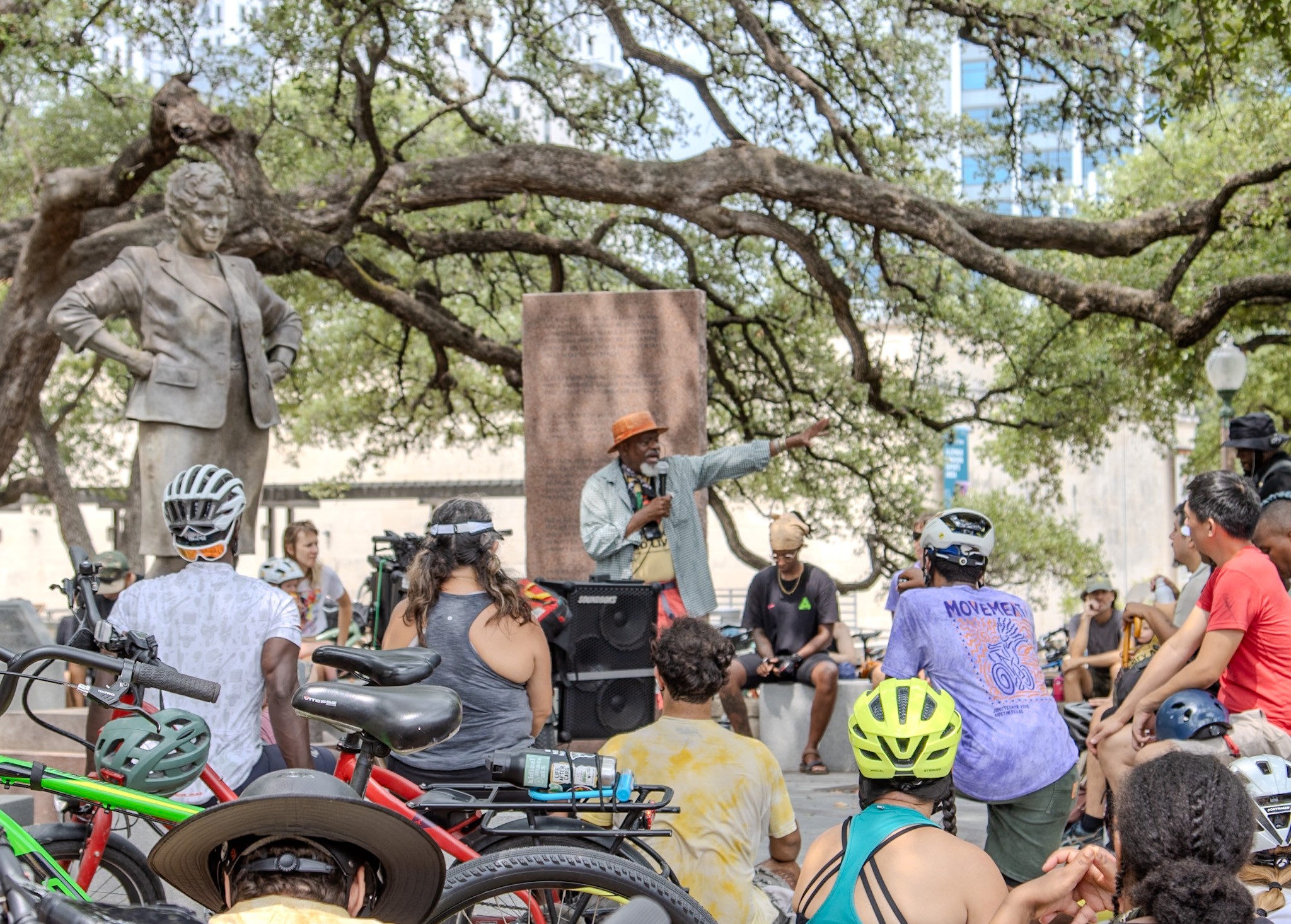 Group of people gathered outdoors around a speaker, with a statue and tree in the background, some wearing helmets.