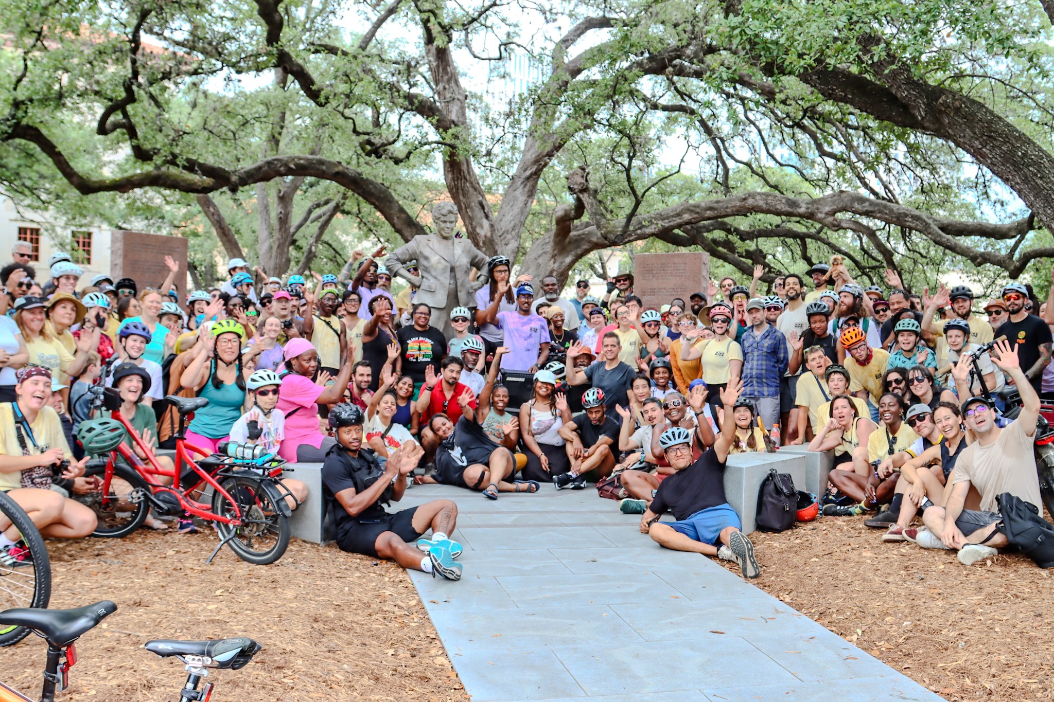 Large group of people gathered outdoors under a sprawling tree, some sitting and some standing, with children and bicycles present.