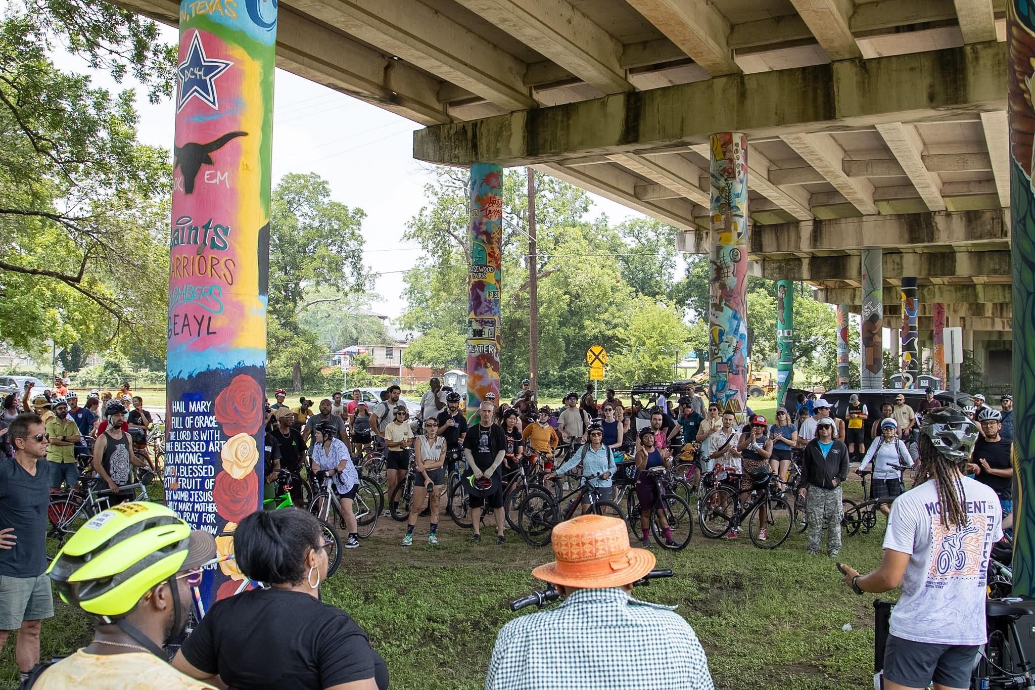Crowd of people gathered under a bridge with colorful pillars, some with bicycles, in a park setting.