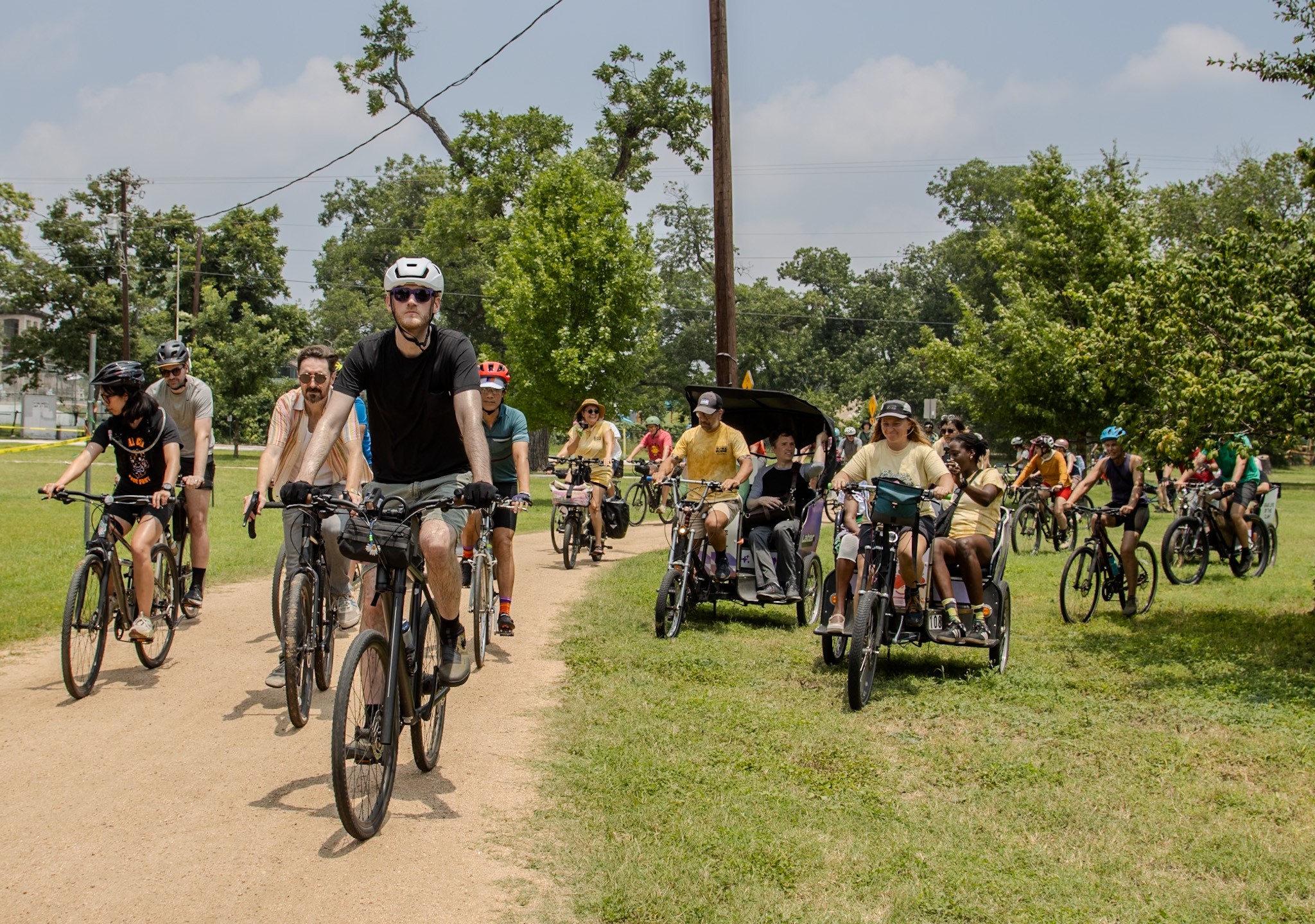 Group of people riding bicycles and using pedal-powered vehicles on a dirt path in a park with trees and a cloudy sky.
