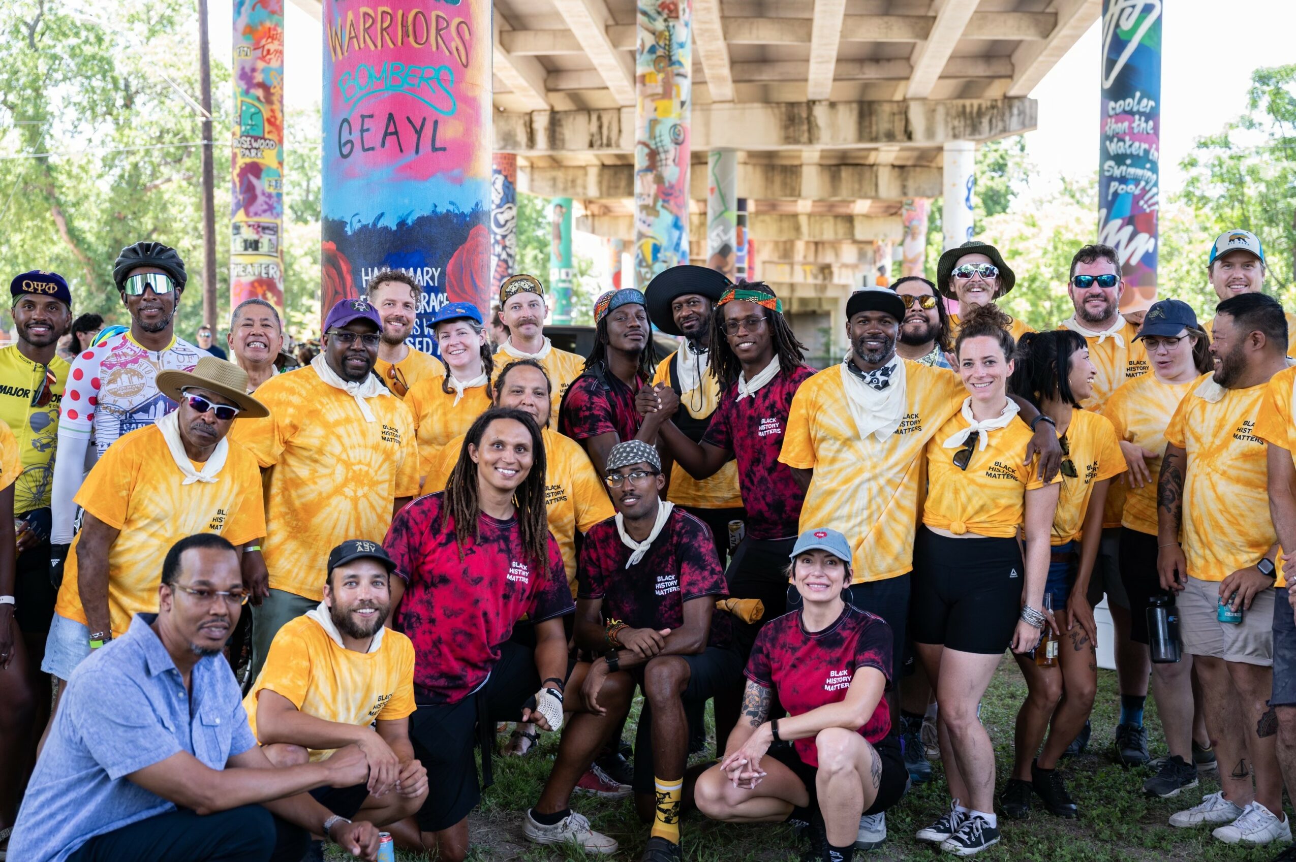 Group of people outdoors under a bridge, some wearing yellow shirts, with colorful banners in the background.
