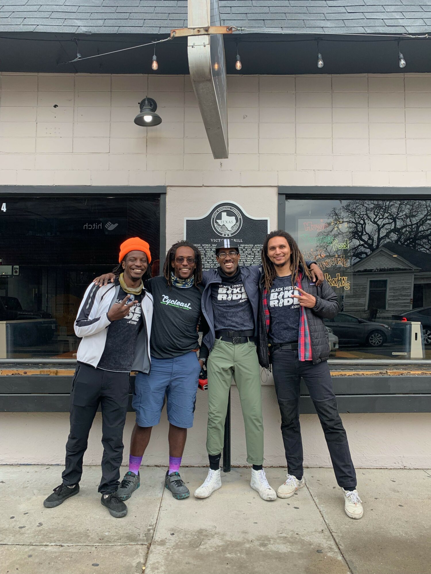 Four people standing outside a building with a logo, posing for a photo, with a window and wall behind them.