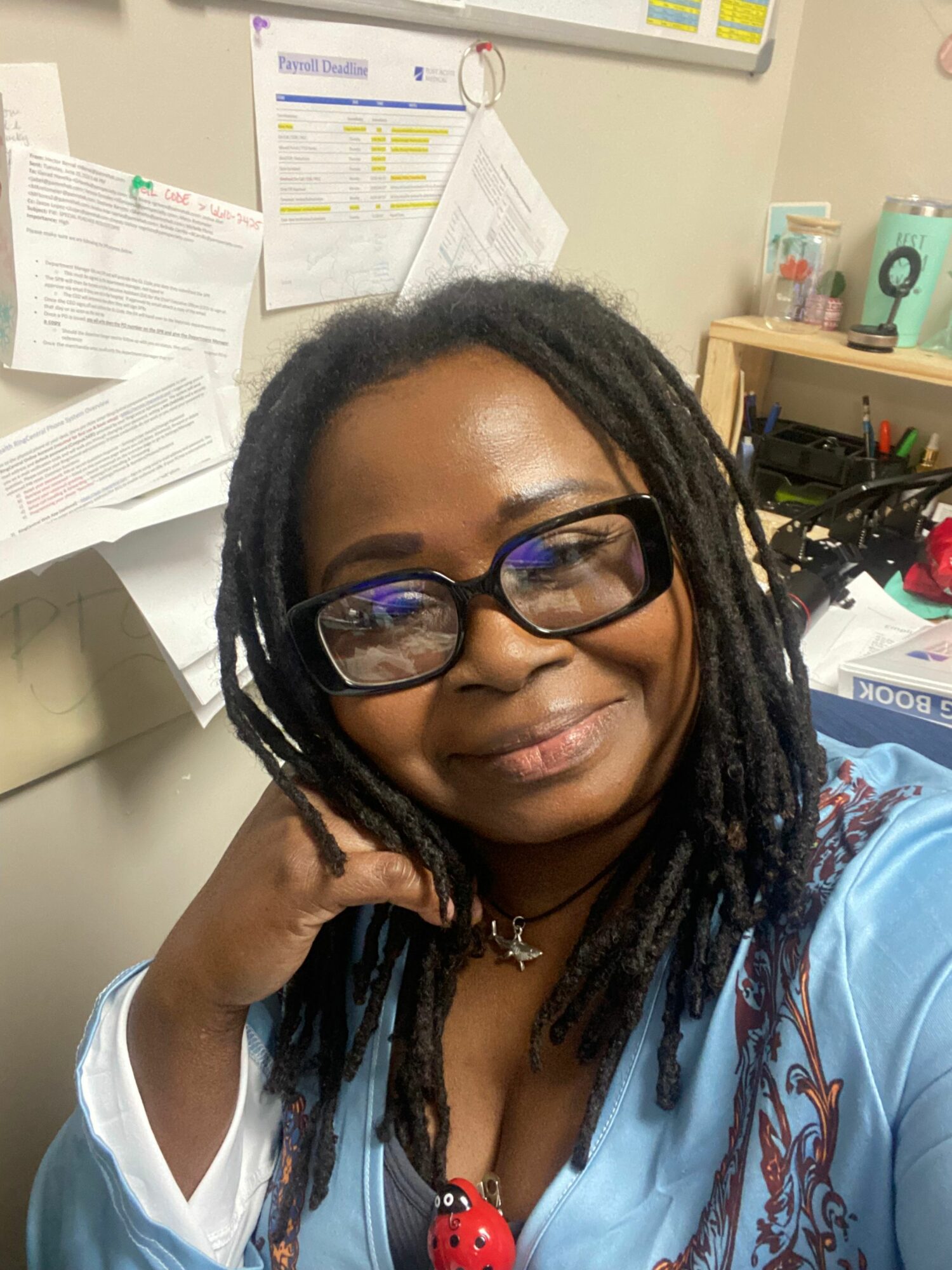 Woman with glasses and dreadlocks smiling, resting her head on her hand in an indoor setting.