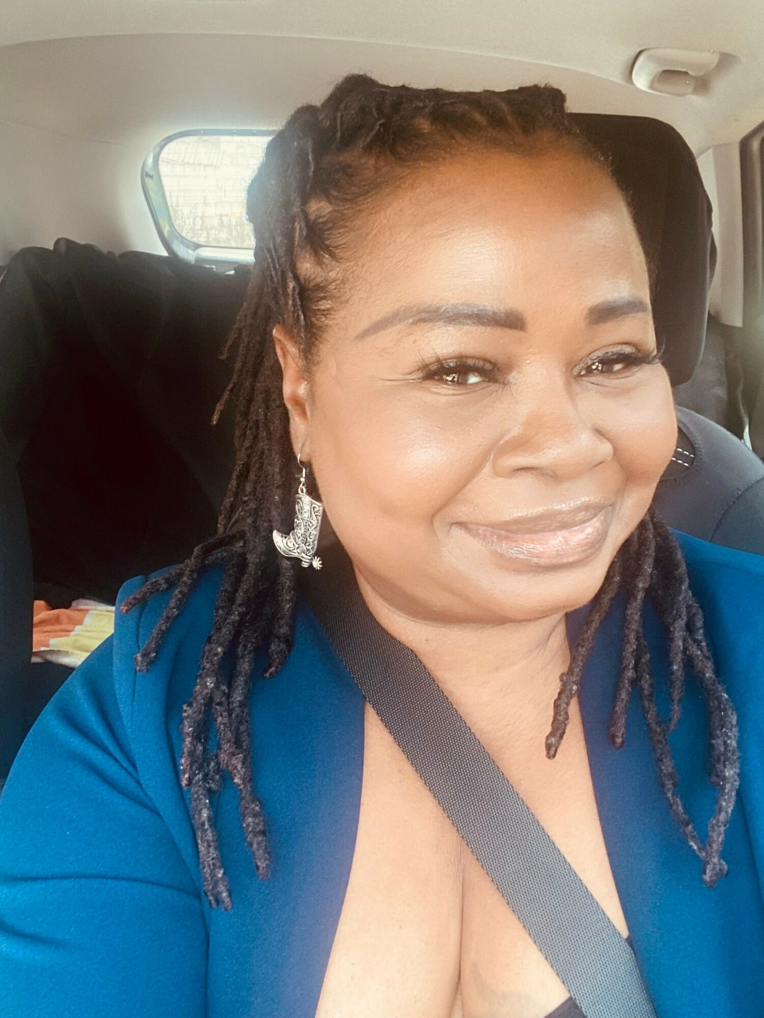 Smiling woman with braided hair wearing earrings and a blue top, sitting in a car.