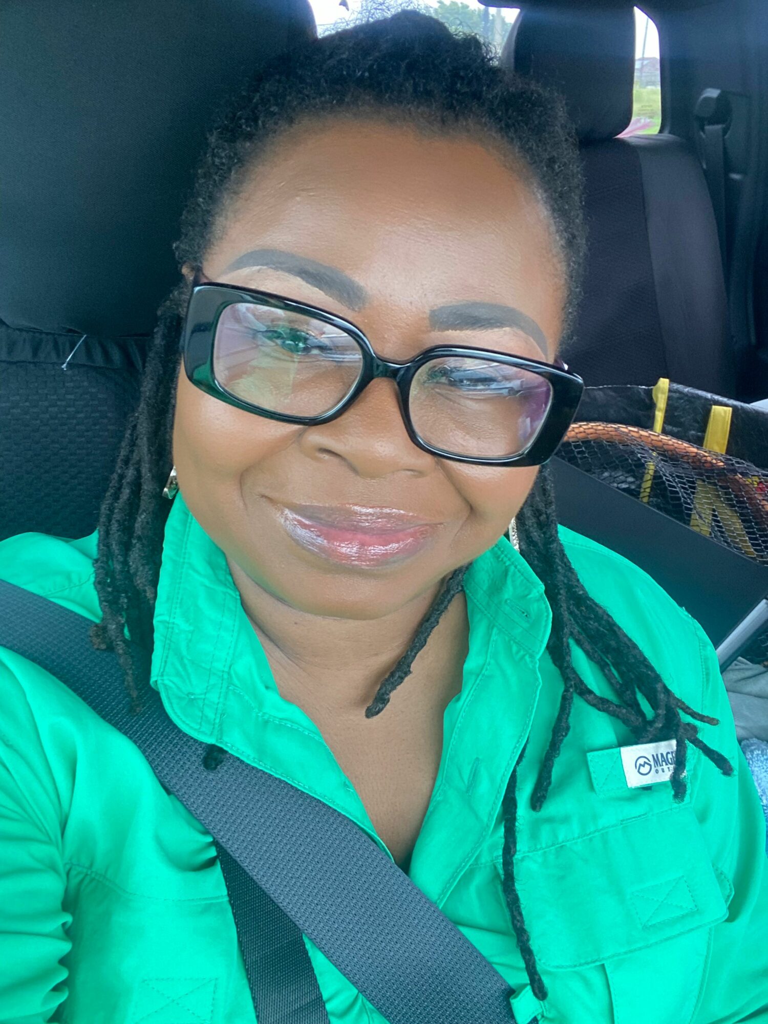 Woman with glasses and braided hair smiling, wearing a green jacket, sitting in a vehicle seat.