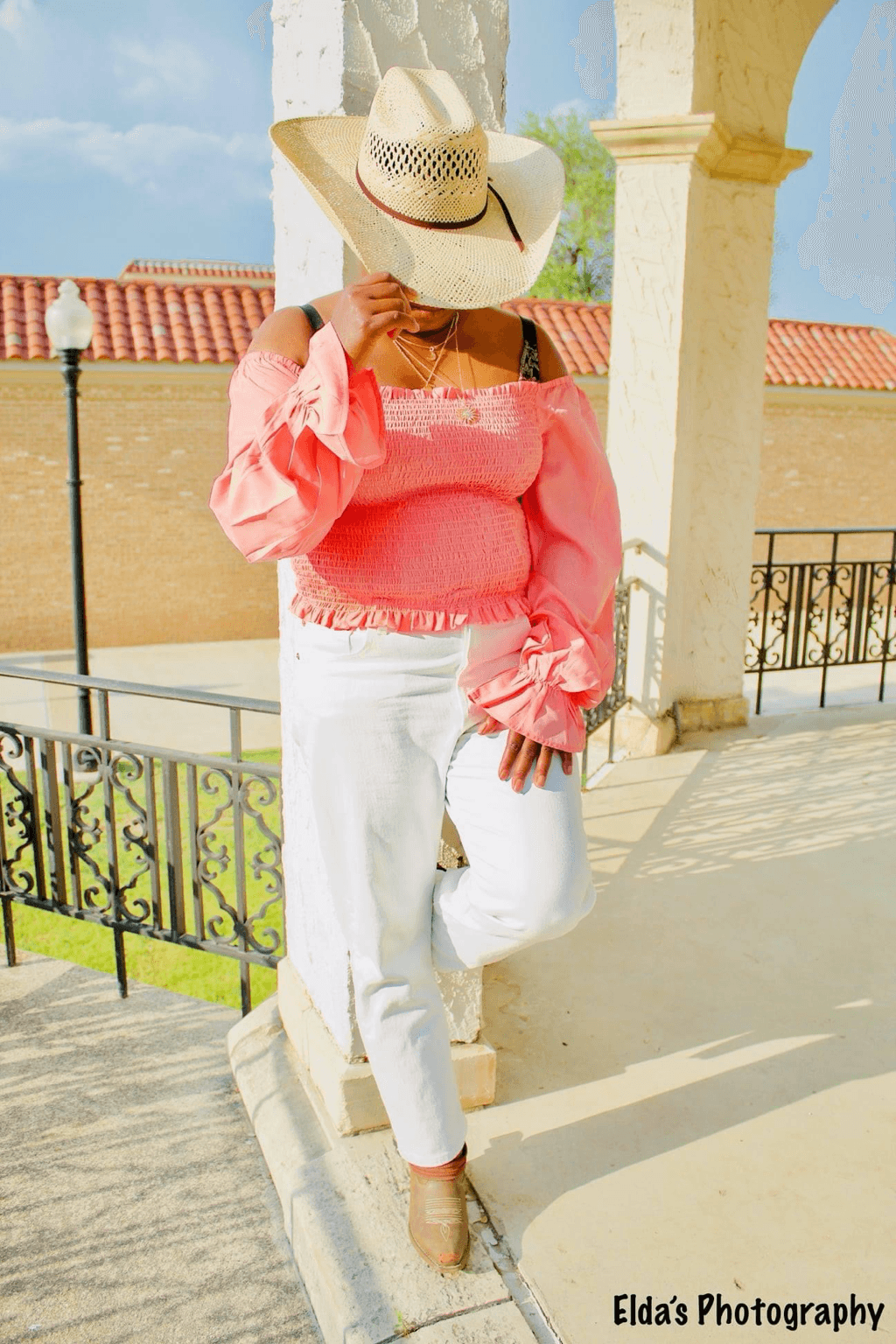 Person wearing a pink off-shoulder top, white pants, and a wide-brimmed hat, standing outdoors near columns and a railing.