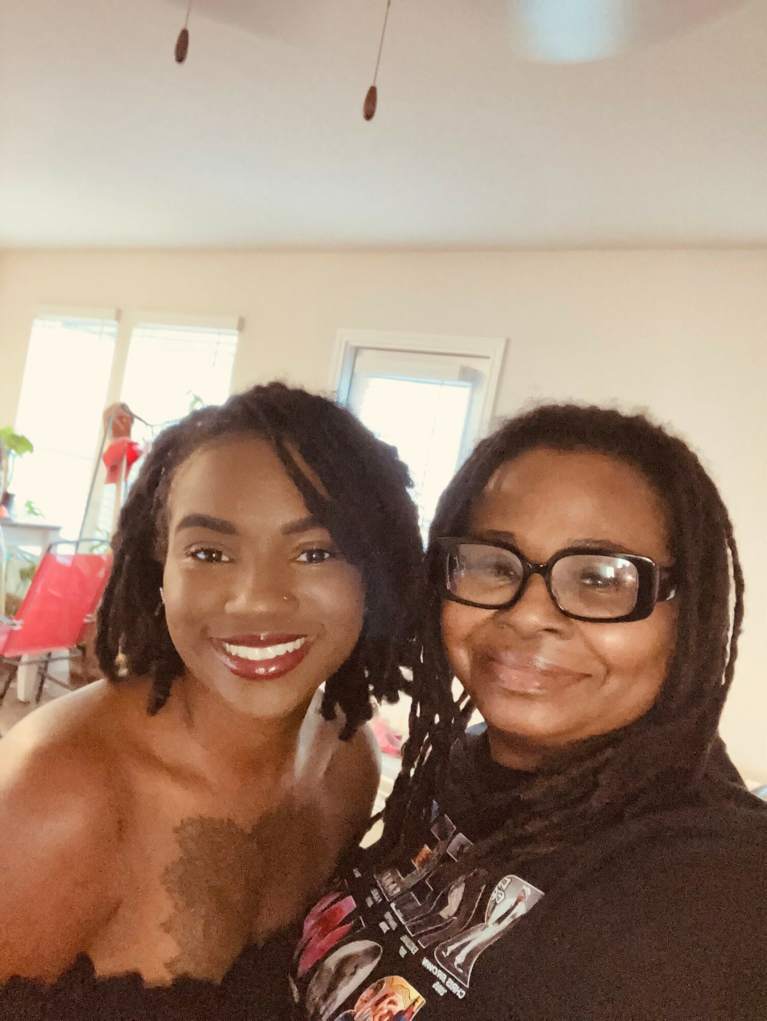 Two women smiling indoors, one with curly hair and glasses, the other with shoulder-length hair, in a bright room.