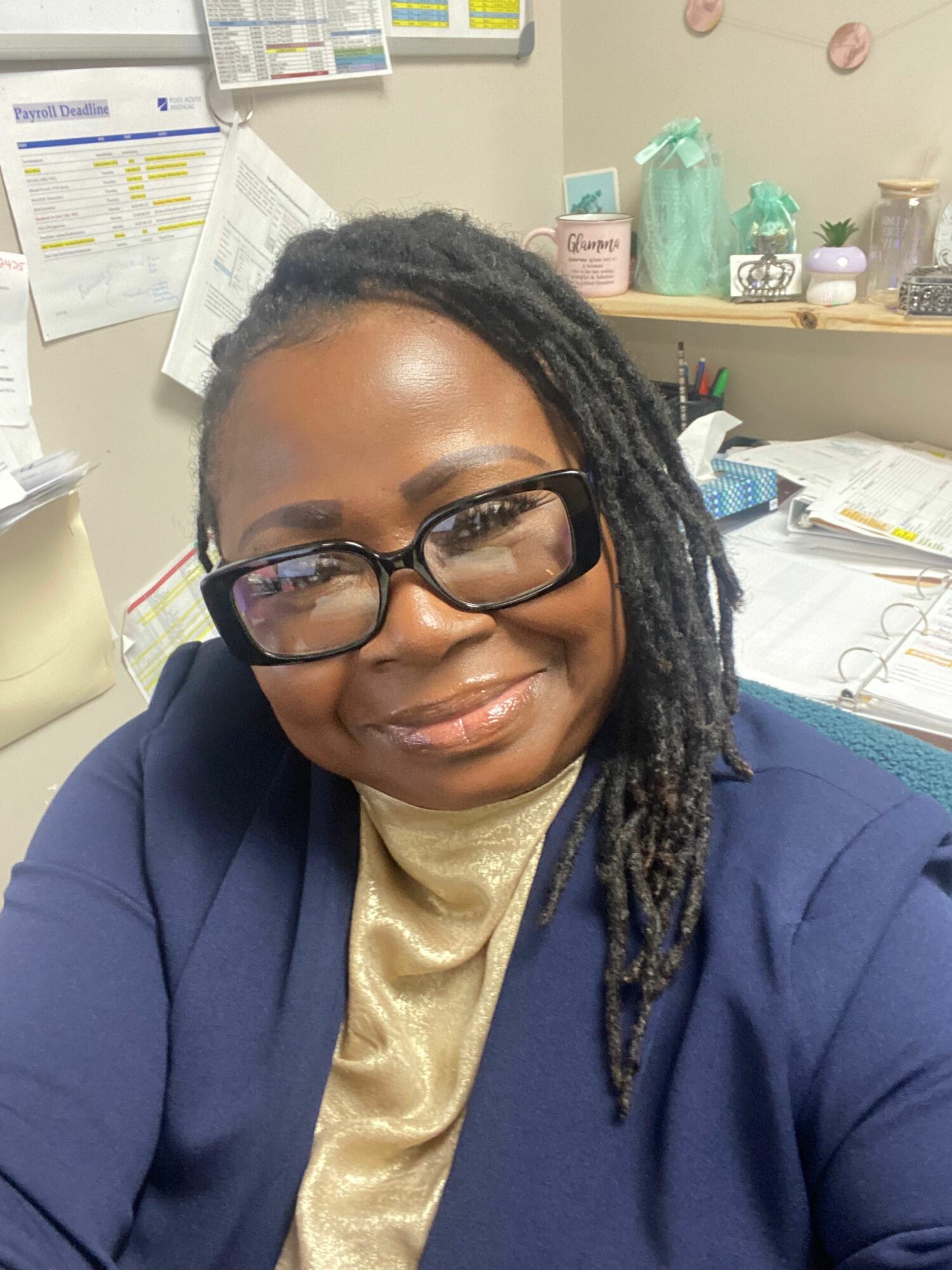 Person with glasses and dreadlocks smiling, wearing a navy blazer and gold scarf, in an office setting.
