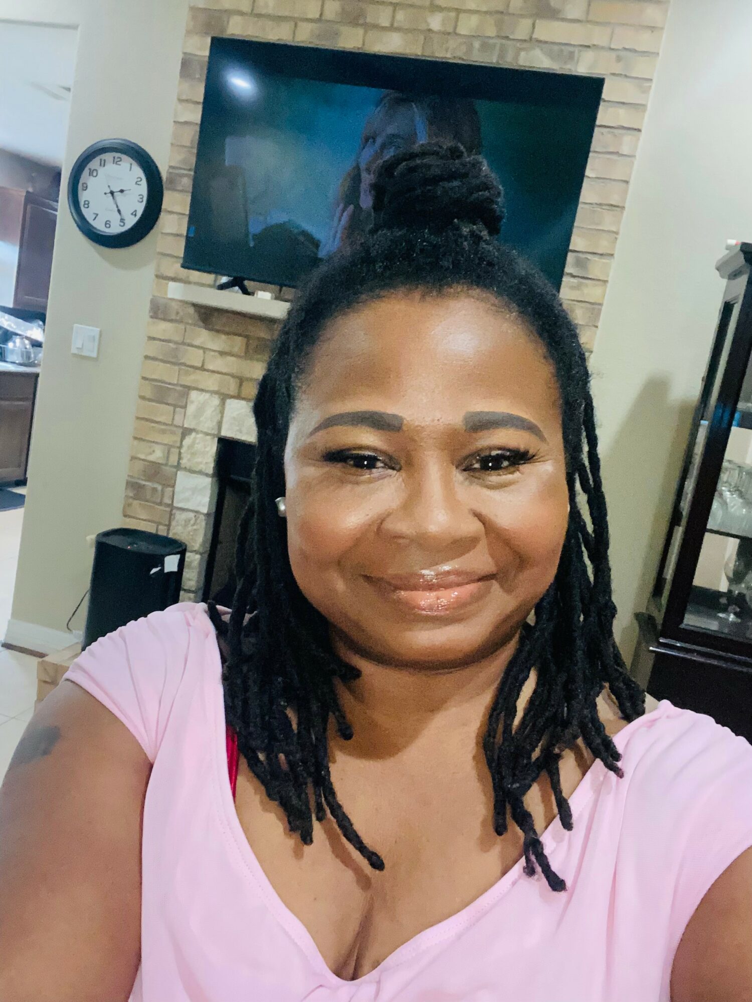 Smiling woman with dreadlocks wearing a pink top, in a room with a TV, clock, and brick wall background.