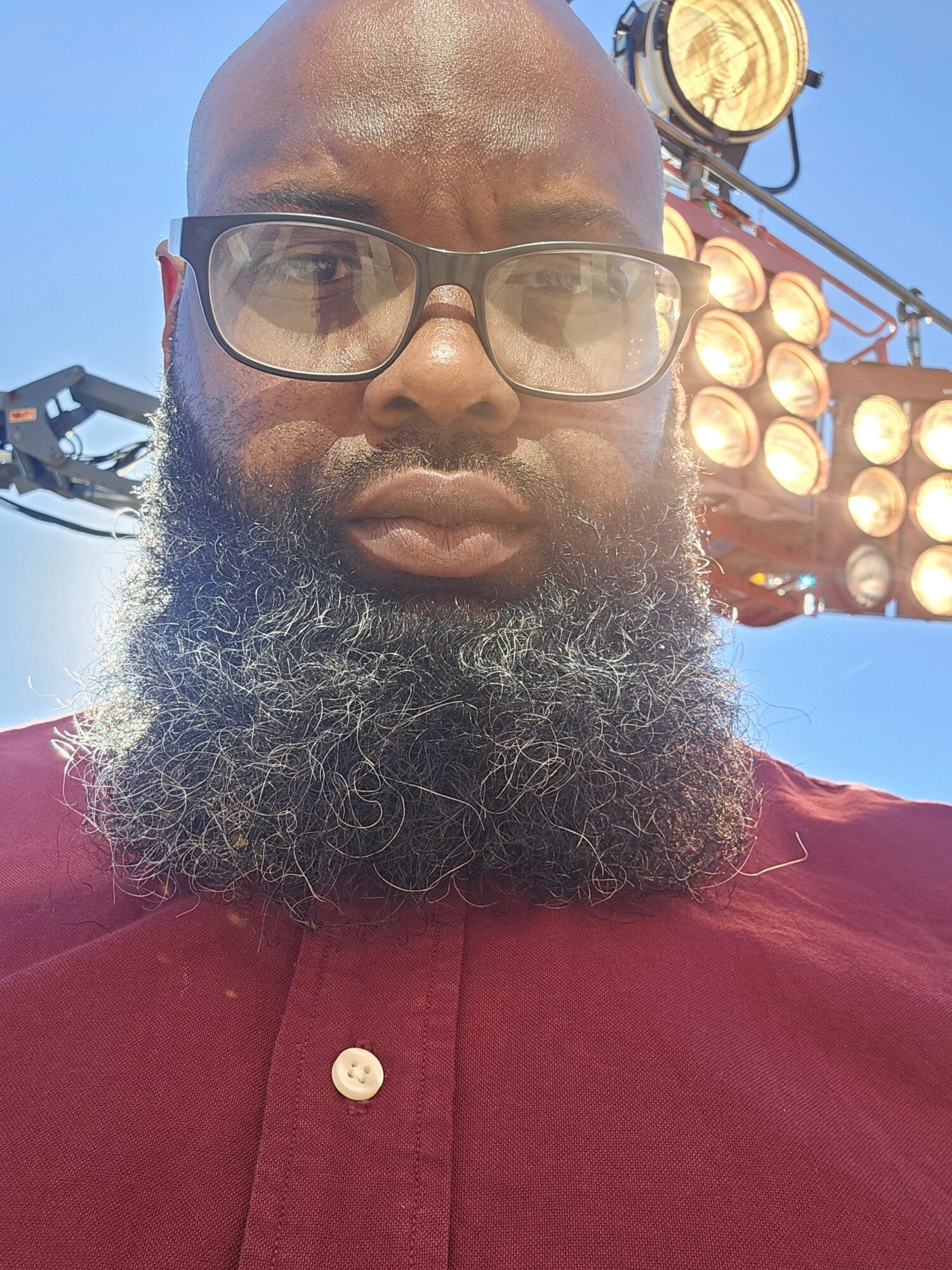 Close-up of a person with glasses and a beard, wearing a red shirt, outdoors with a blue sky and a person in the background.