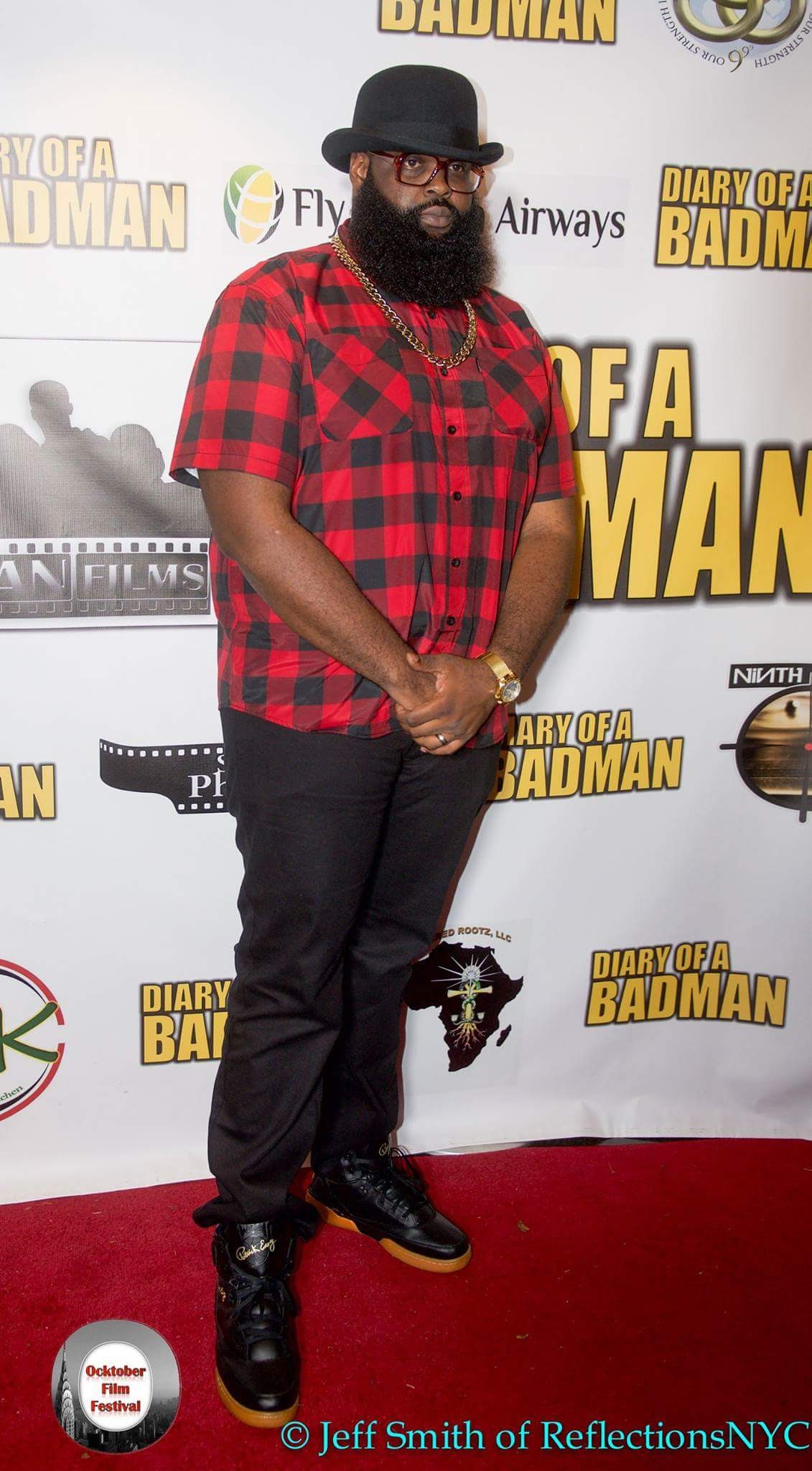 Person with a beard and glasses wearing a red plaid shirt, black pants, and boots, standing on a red carpet.