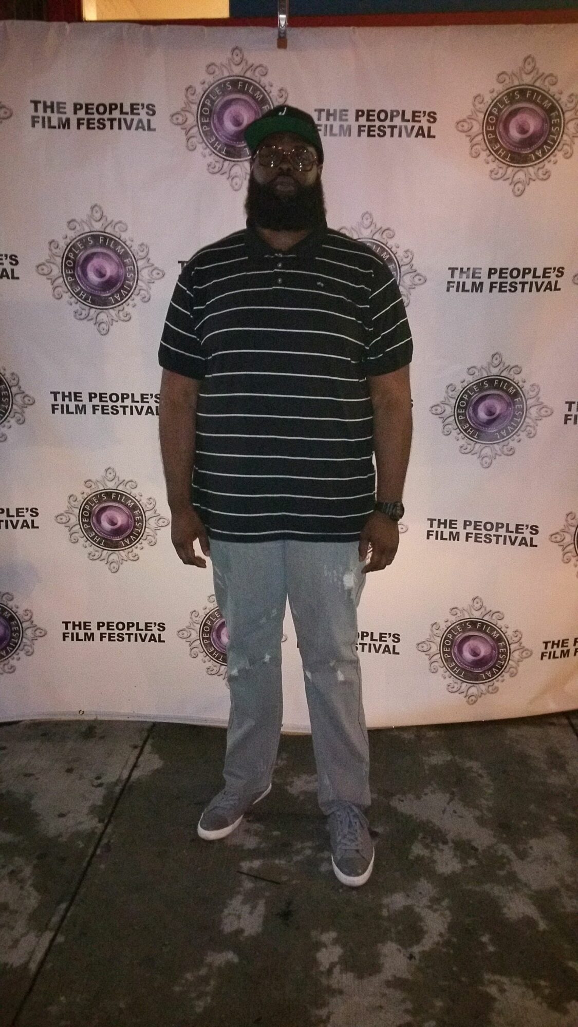 Person standing in front of a backdrop with logos, wearing a striped shirt, jeans, and a face mask.