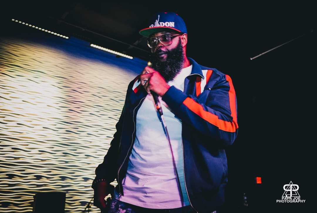 Person with a beard, sunglasses, and a cap holding a microphone, standing in front of a water background.