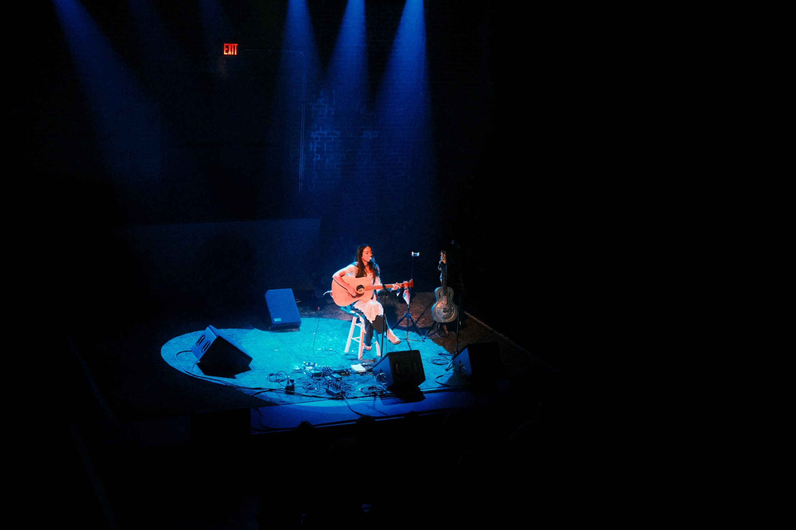 Musician playing guitar on stage with blue lighting, surrounded by equipment, in a dark theater setting.