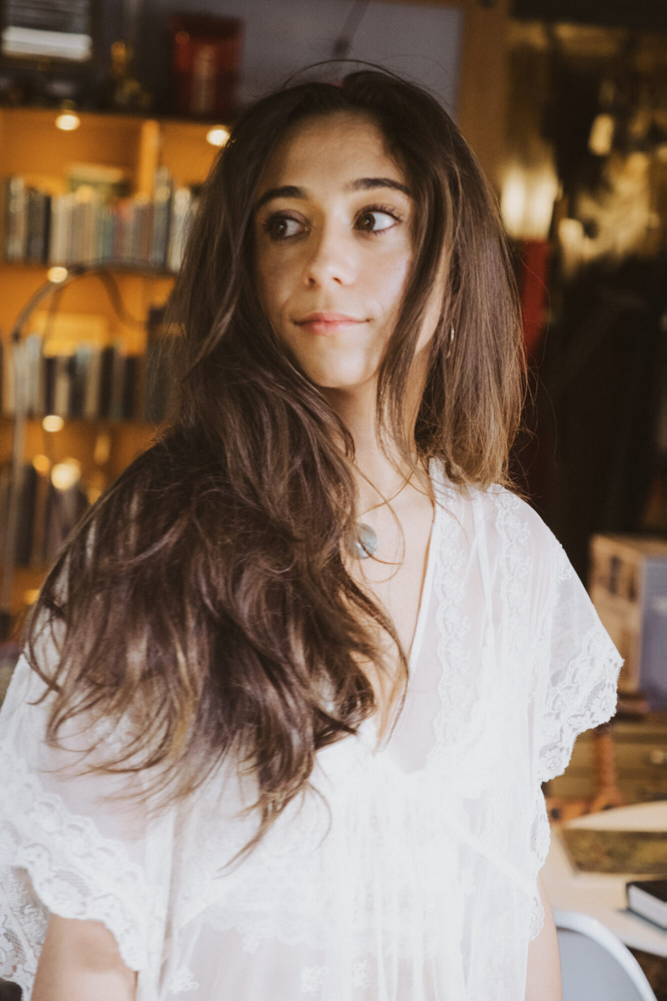 Young woman with long, wavy hair wearing a white top, looking to the side in a warmly lit indoor setting.