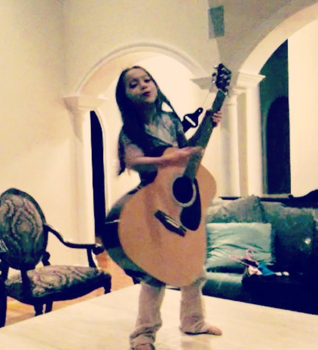 Girl standing indoors holding an acoustic guitar, smiling, with furniture and a sofa in the background.