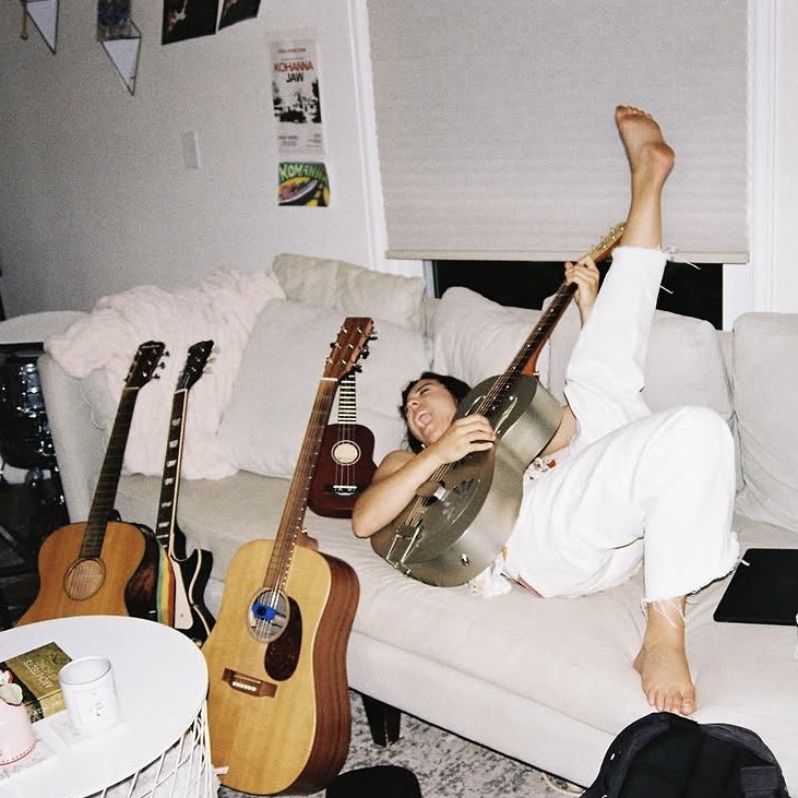 Person lying on a sofa holding an acoustic guitar, with four other guitars nearby, in a living room.