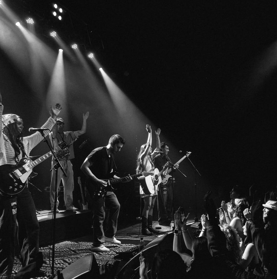 Musicians perform on stage with audience raising hands, stage lights shining down, in black and white photo.