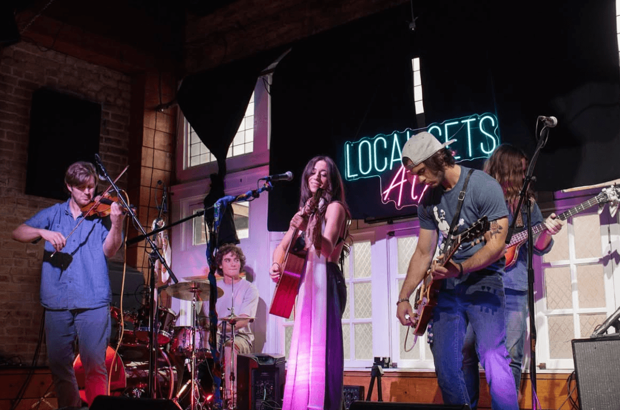 Group of five musicians performing on stage with a neon sign reading 'Local Sets' behind them.