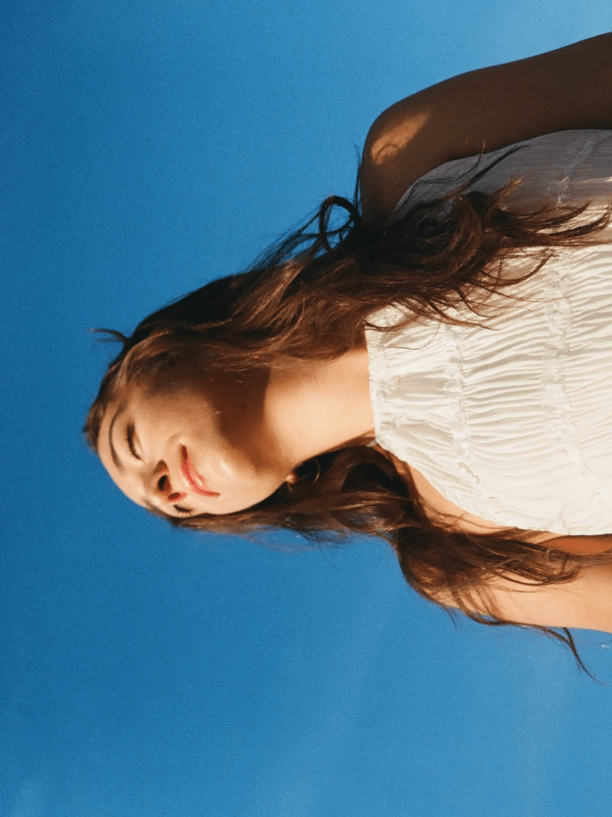 Young woman with long hair and a white top, standing against a blue sky, looking to the side.
