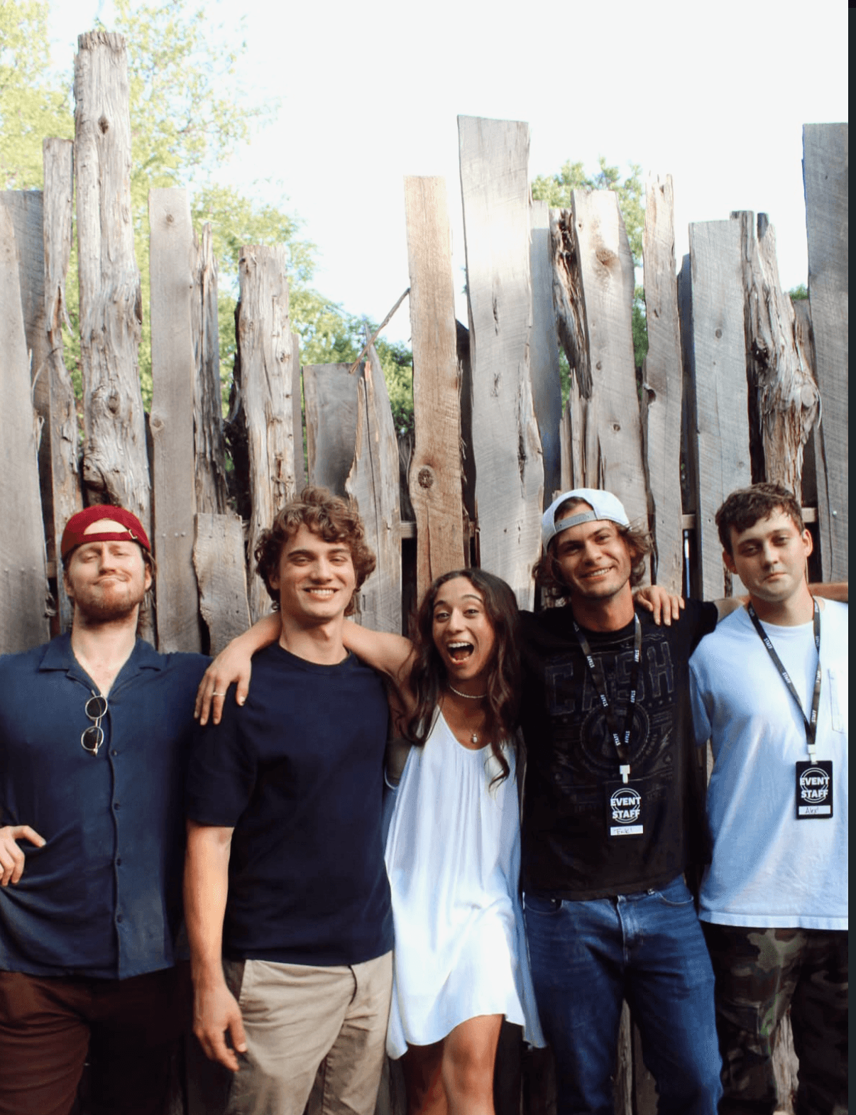Group of five people standing outdoors in front of a wooden fence, smiling, with trees in the background.