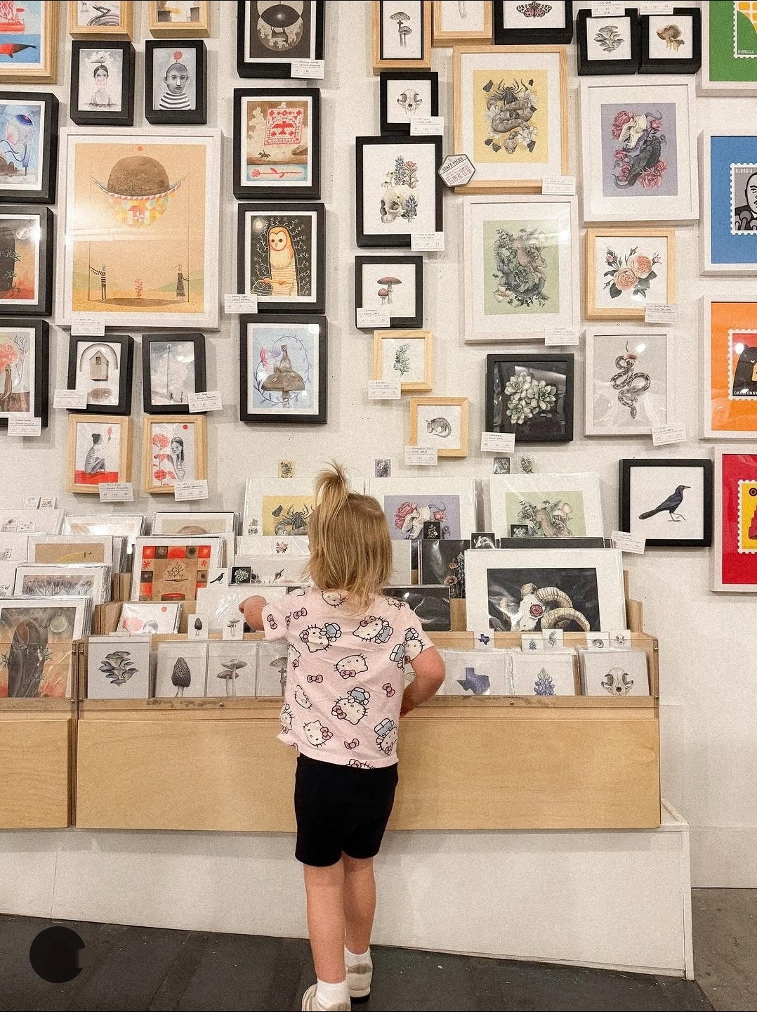 Child with blonde hair wearing a pink shirt and black shorts looks at framed artwork on a gallery wall.