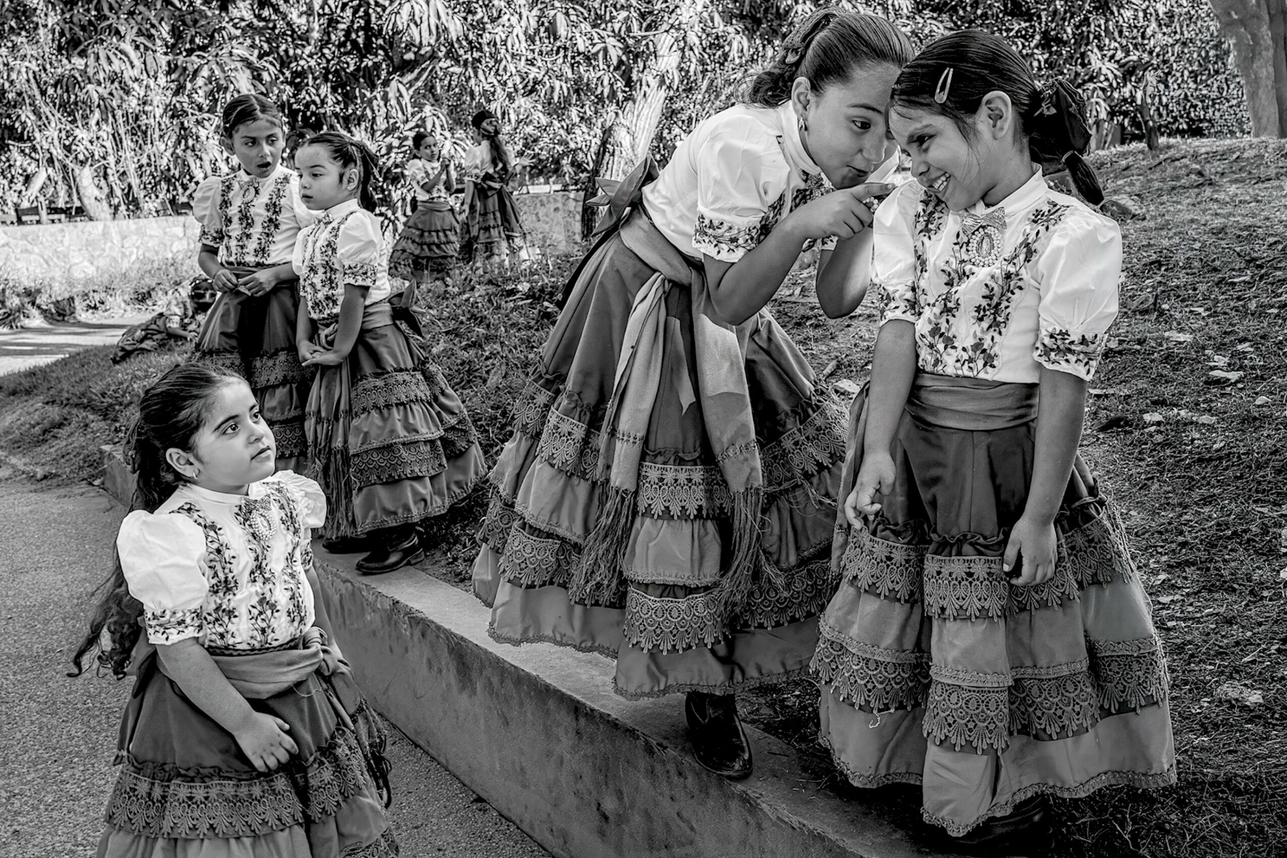 Group of girls in traditional dresses outdoors, some standing on a low wall, others sitting nearby, trees in background.