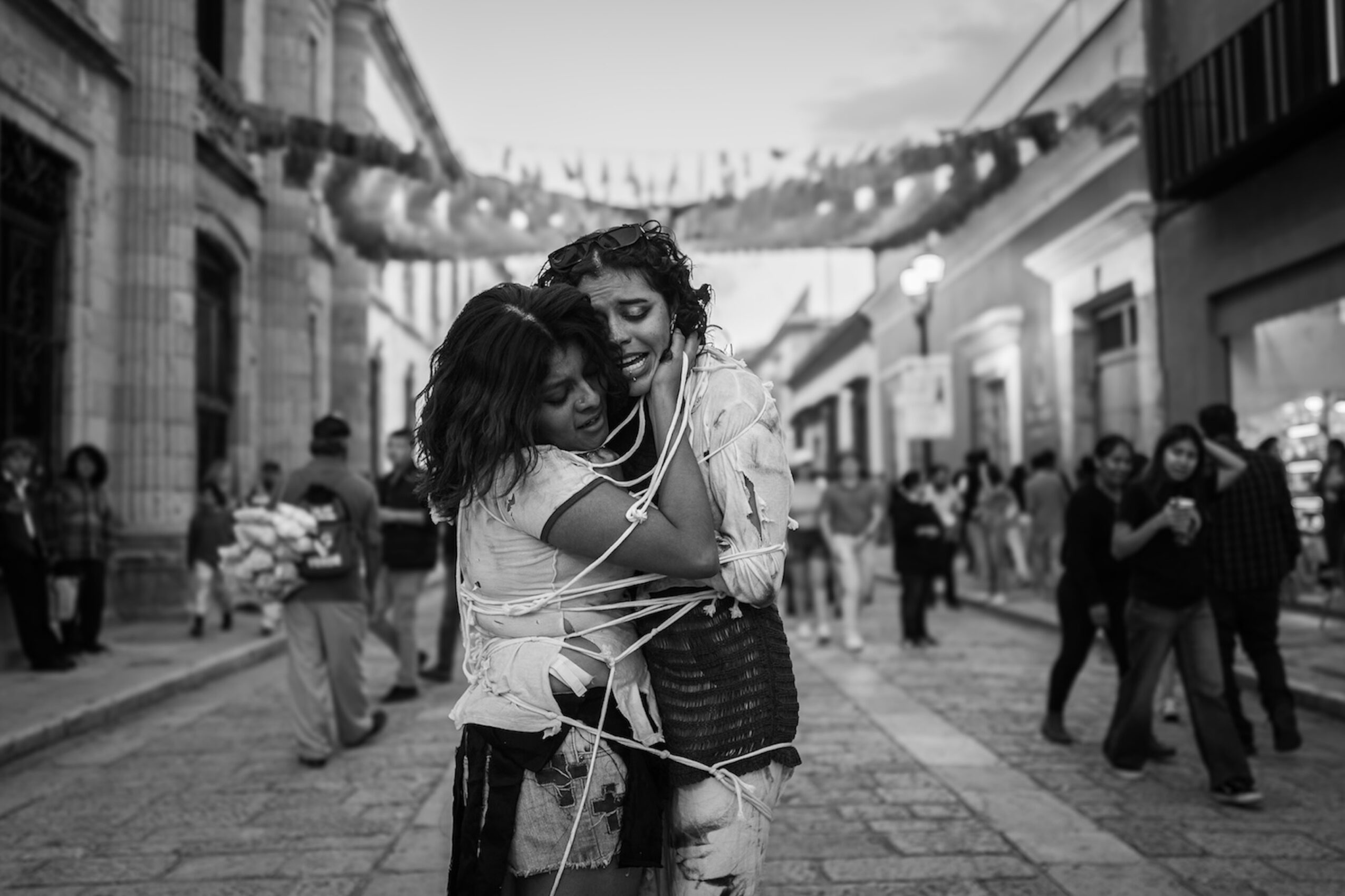 Two women embrace and smile in a busy outdoor shopping street with people walking and buildings in the background.