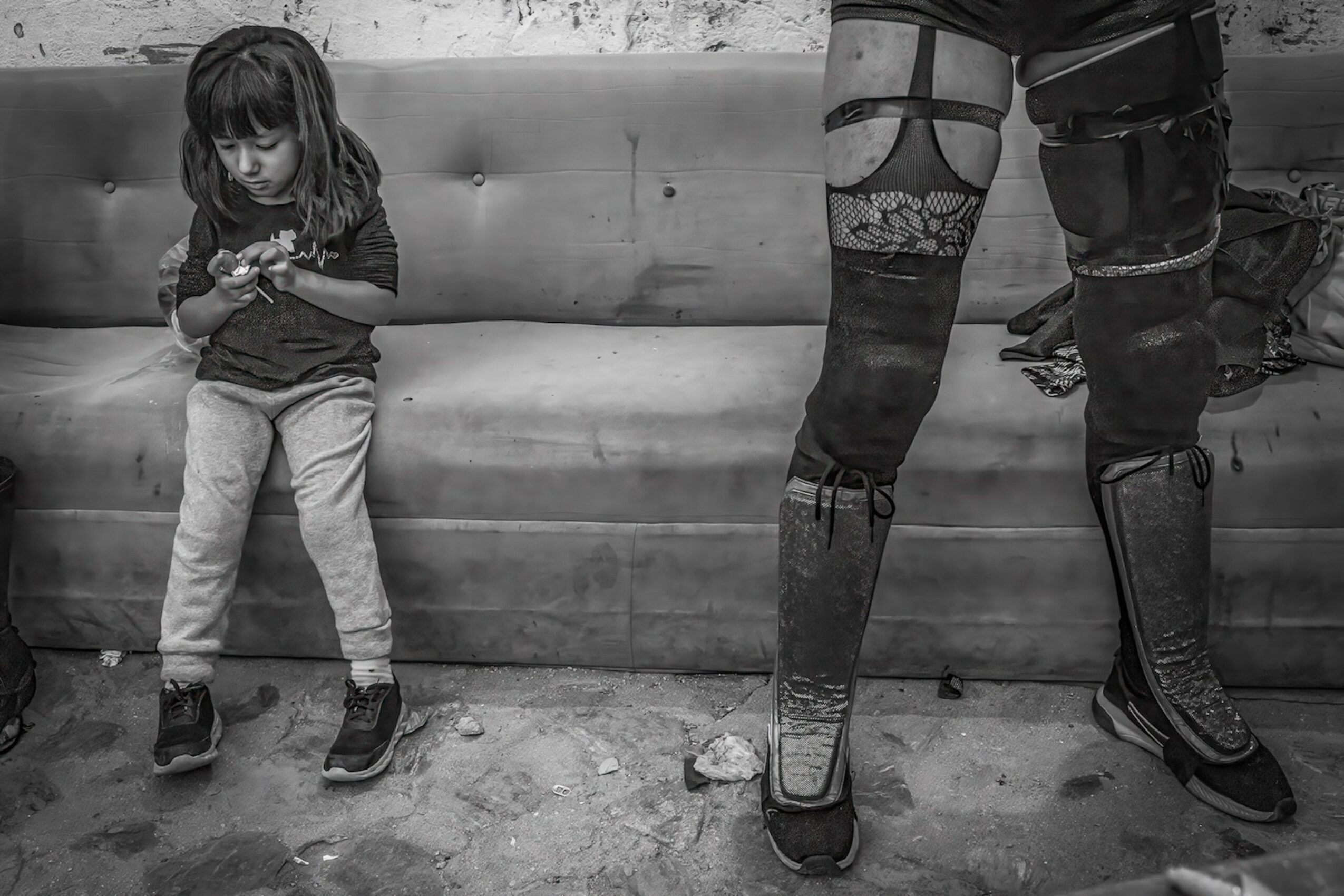 Child sitting on a sofa looking at a device, with a person standing nearby wearing patterned stockings and boots, in black and white.