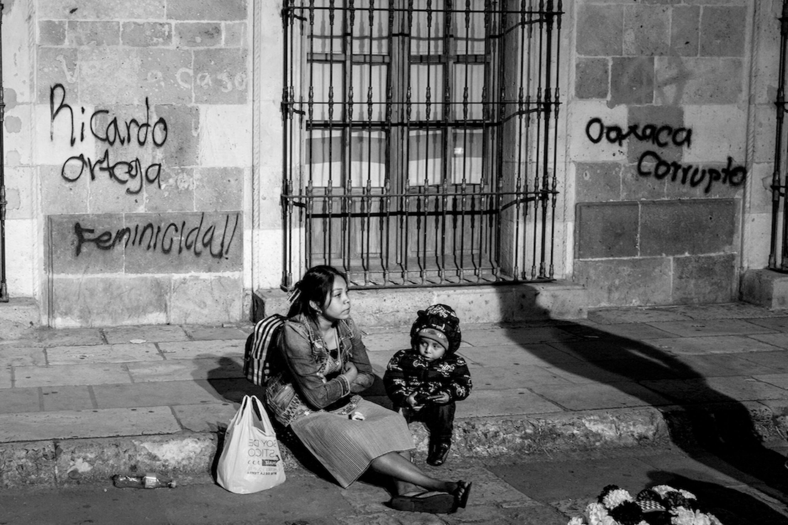Woman and child sitting on sidewalk near a barred window, graffiti on wall, shadow cast on ground, bag beside woman.