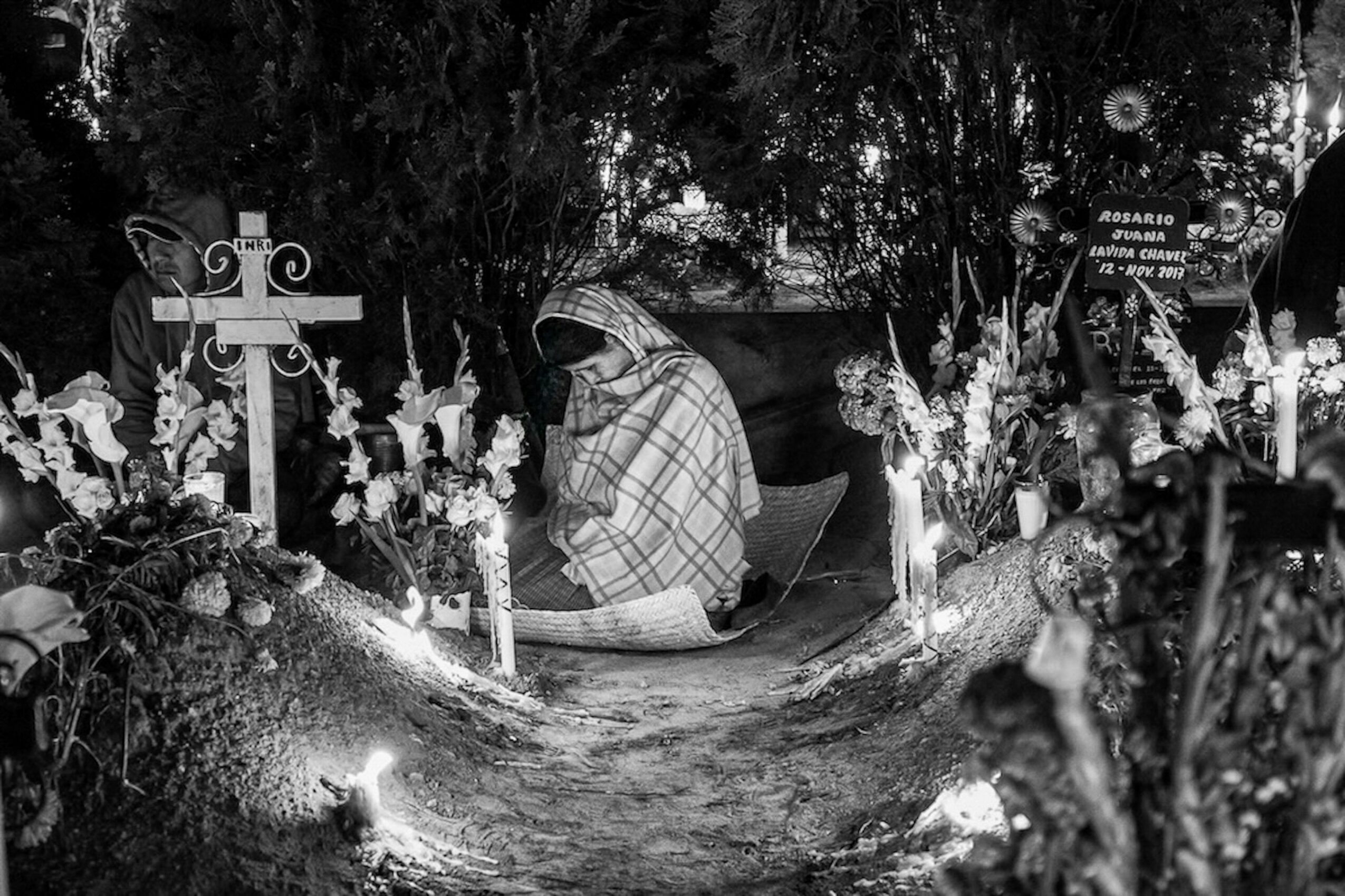Person kneeling at a gravesite with flowers and crosses, holding a hand to their face, in a cemetery.