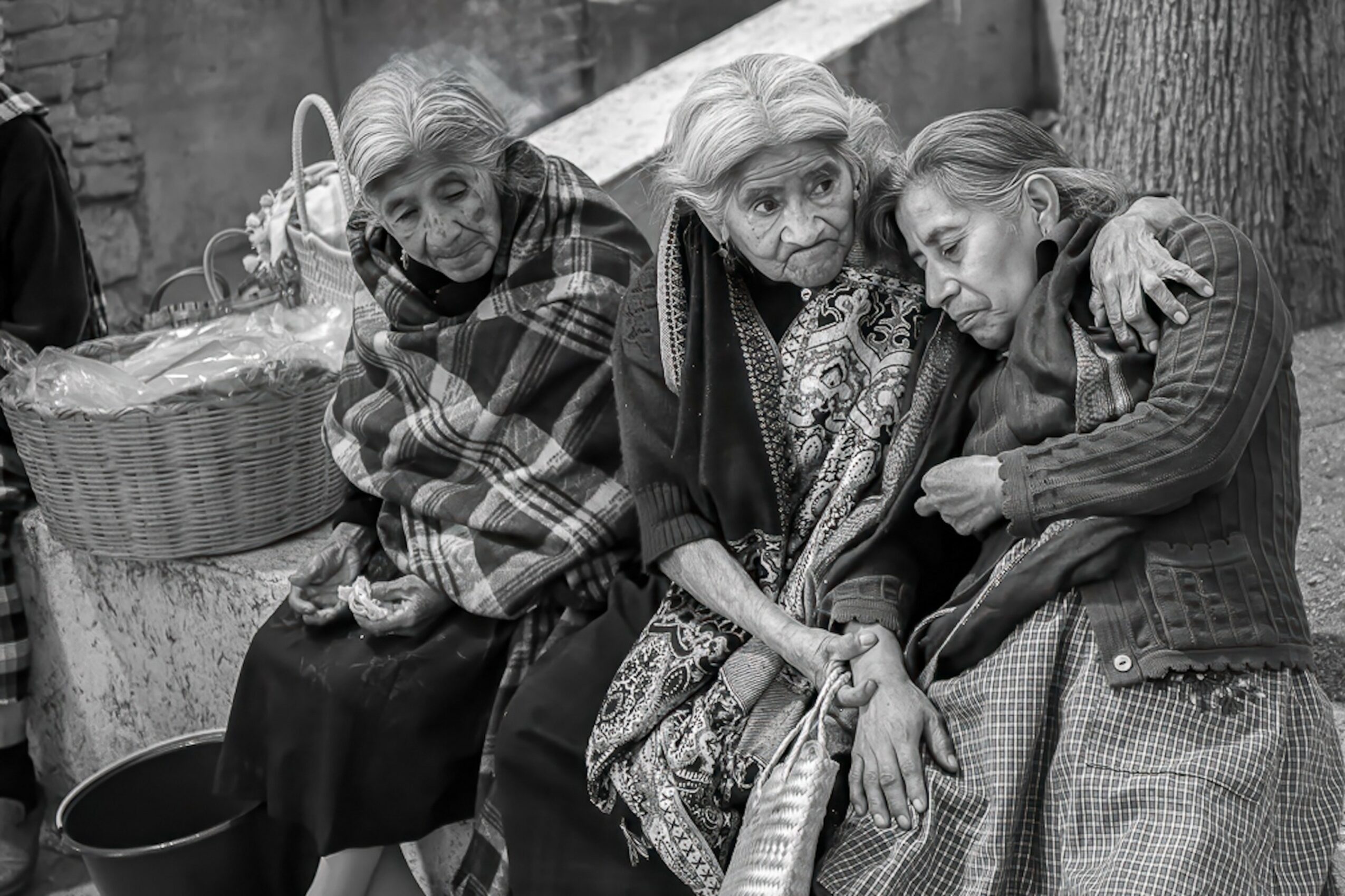 Three women sitting outdoors, one with a basket, two embracing, in black and white.