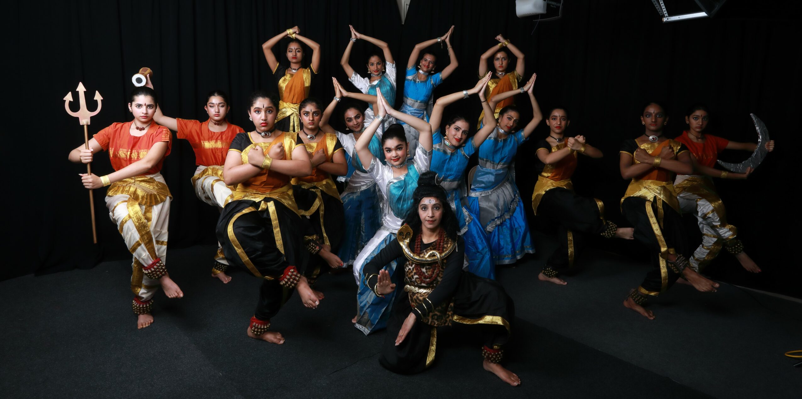Group of dancers in colorful costumes performing on stage with black background.