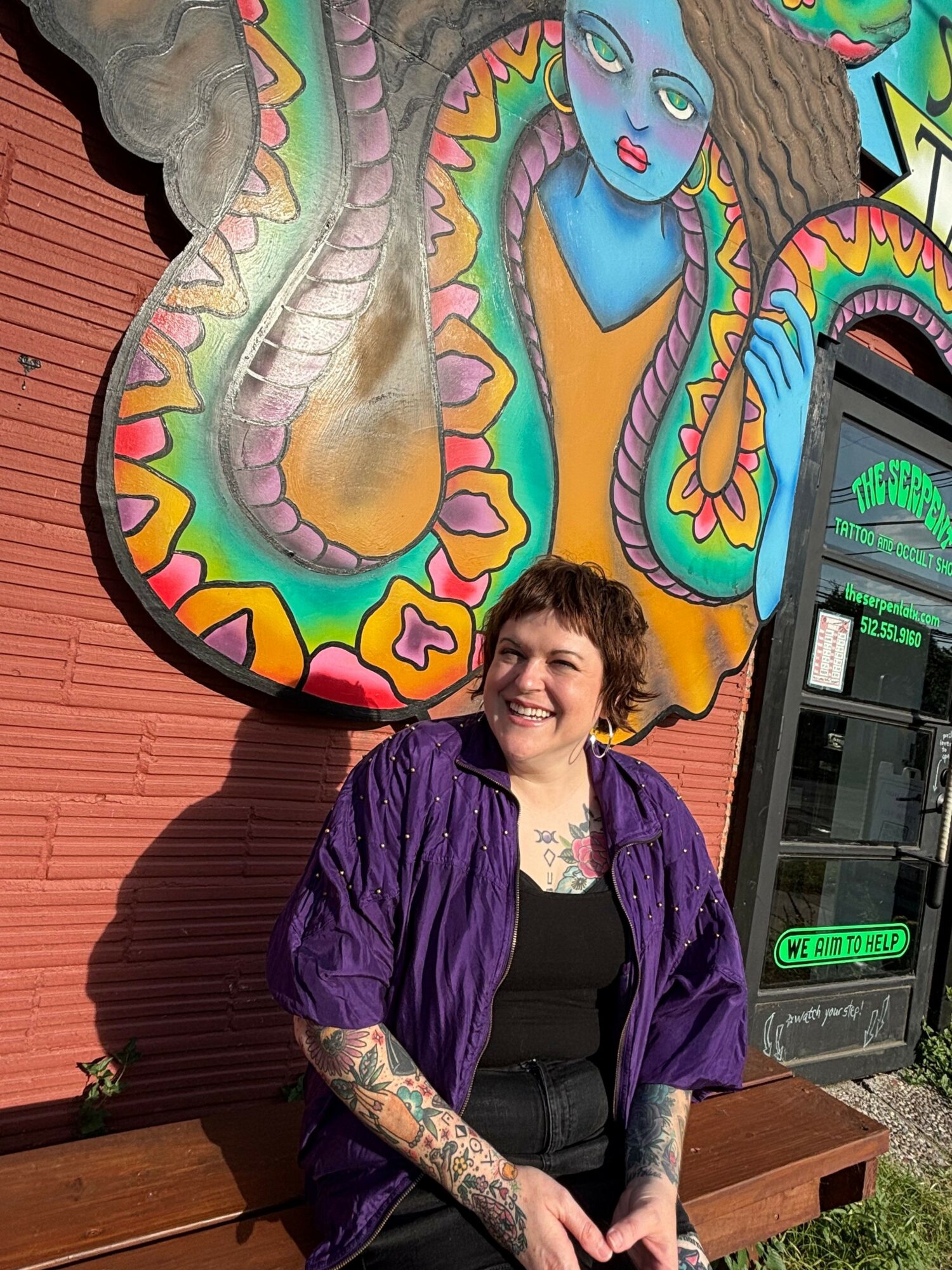 Woman with tattoos and purple jacket lying on wooden deck near colorful snake mural, smiling at camera.