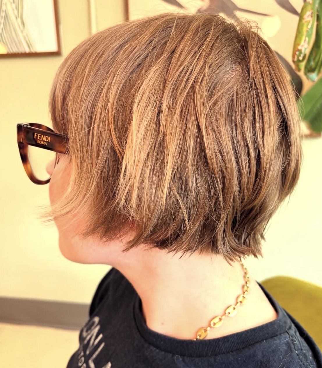 Side view of a person with short, layered, light brown hair, wearing glasses and a black shirt, with a gold necklace.
