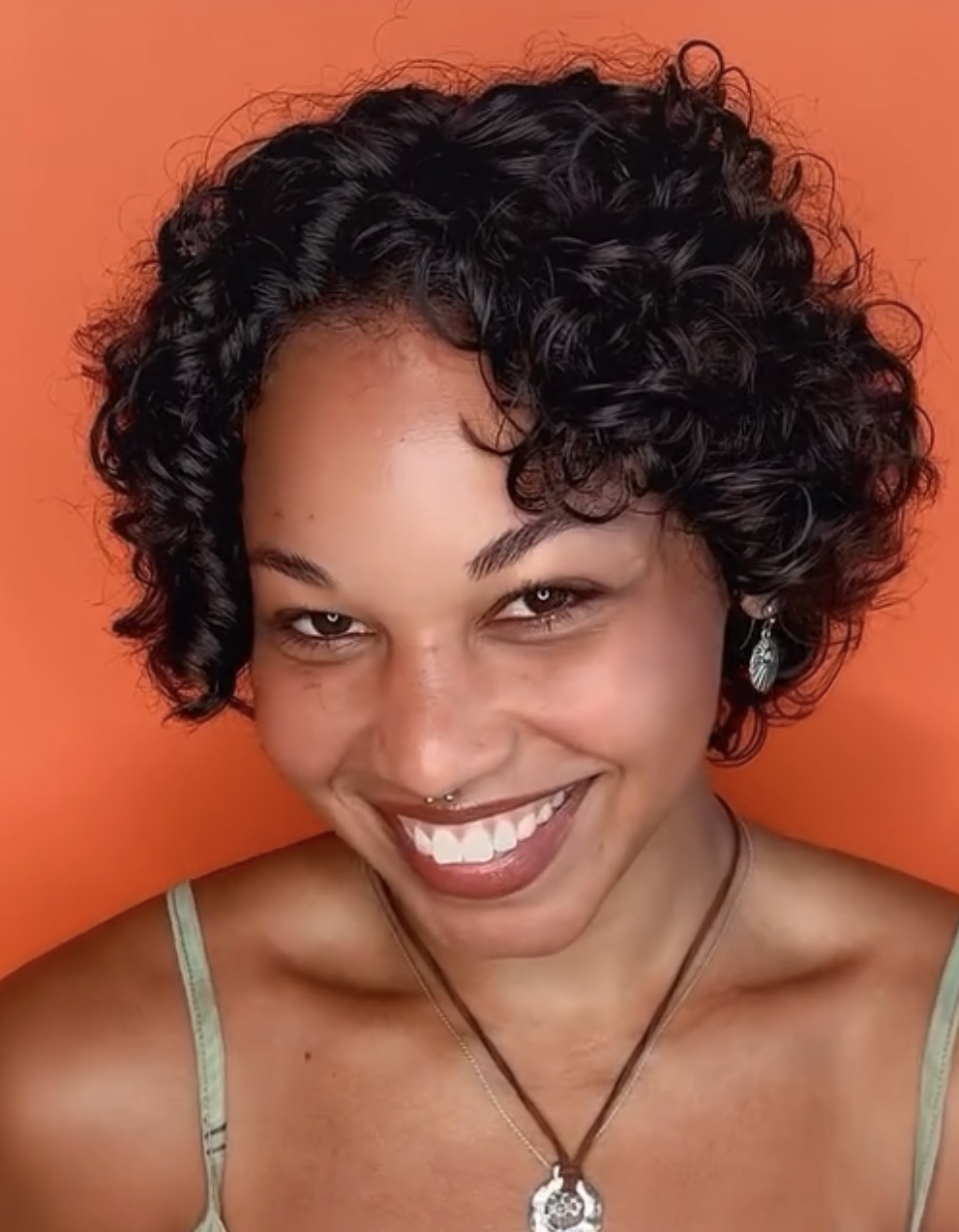 Smiling woman with curly hair, wearing earrings and a necklace, against an orange background.