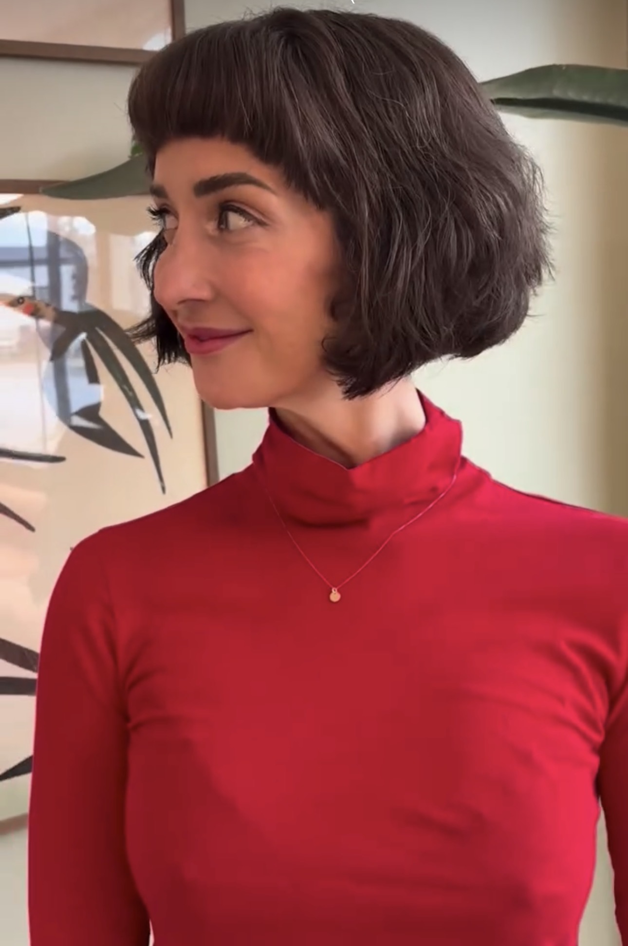 Woman with short dark hair wearing a red turtleneck, looking to the side indoors.