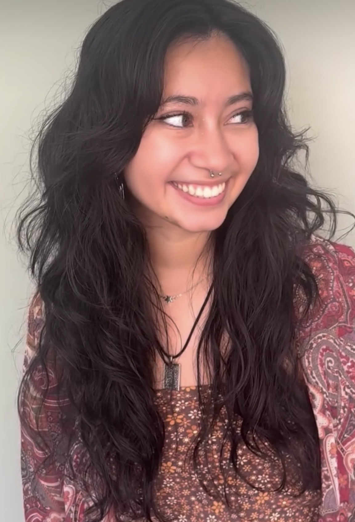 Young woman with long, wavy dark hair smiling and looking to the side, wearing a patterned top and necklace.