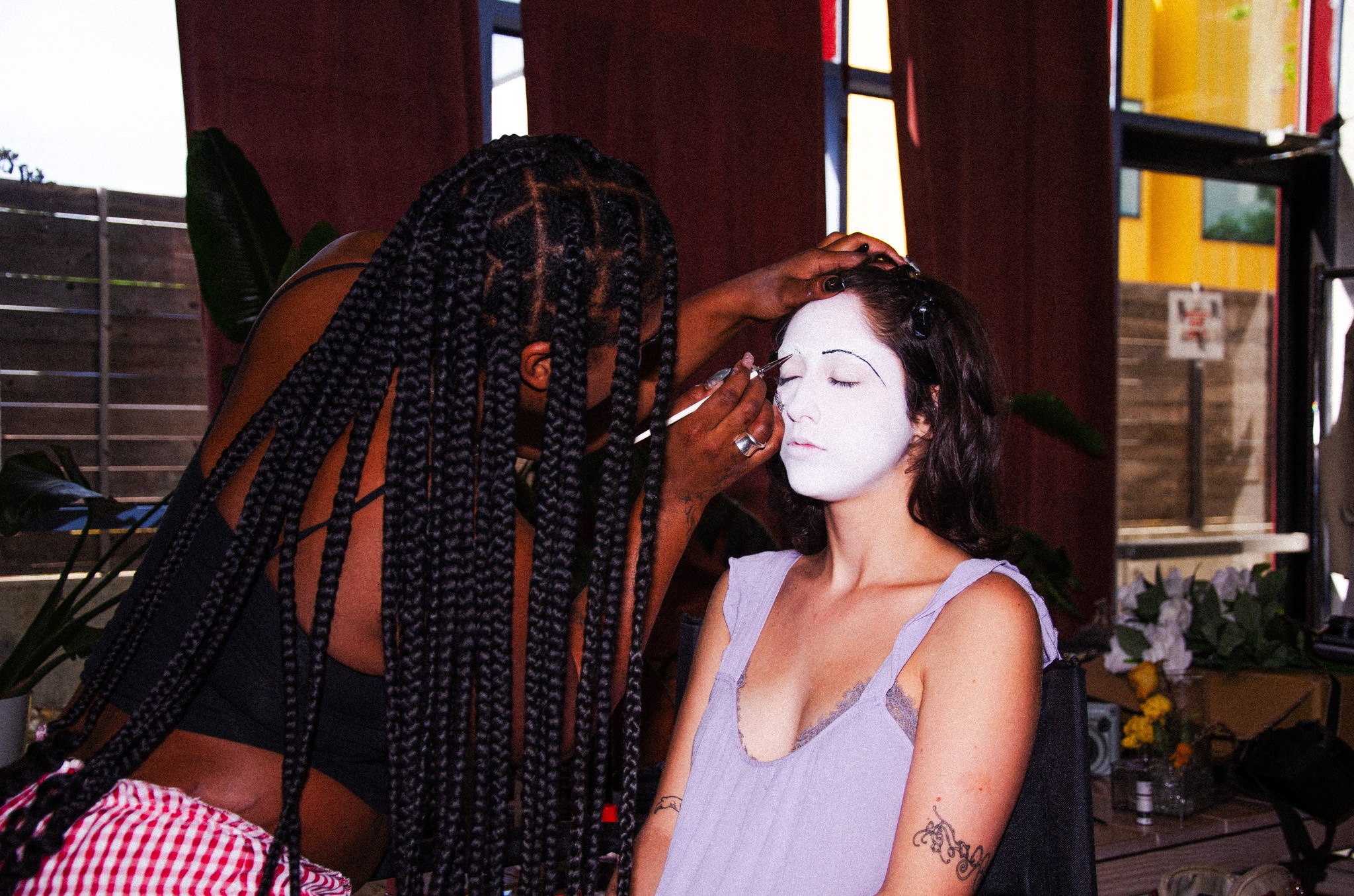 Makeup artist applies white face paint to a woman with dark hair in a room with red curtains and plants.