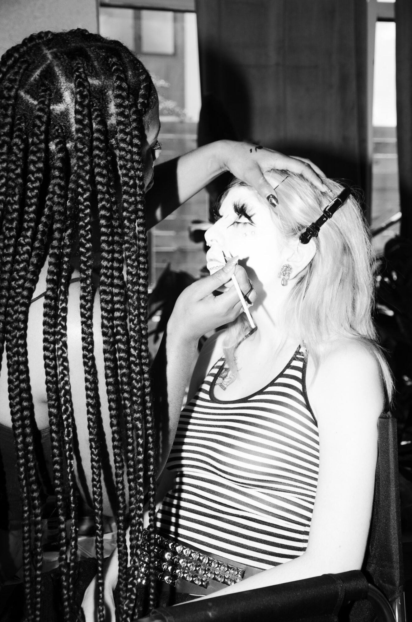 Person with braided hair adjusts glasses on a smiling woman wearing a striped top, indoors with windows in background.