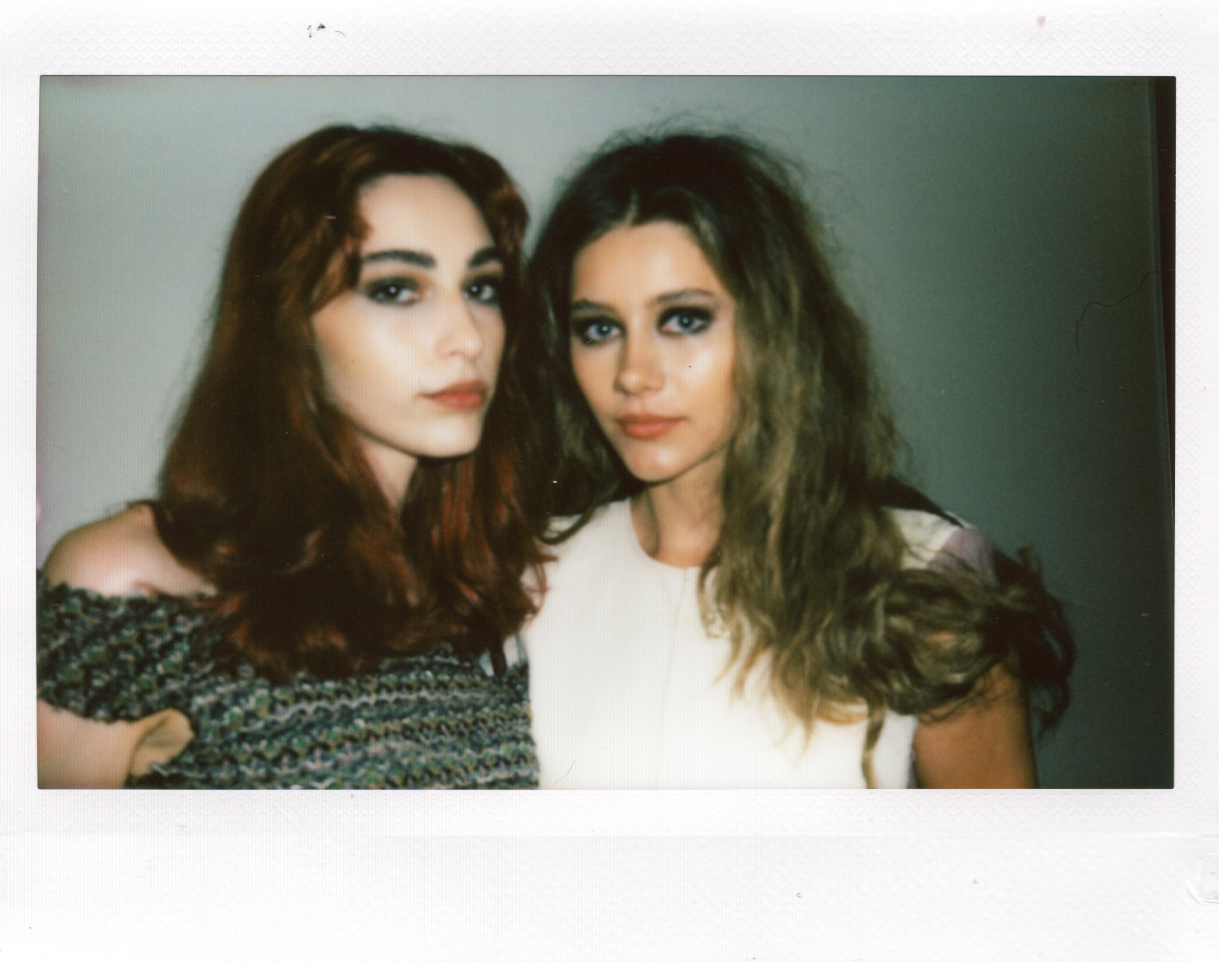 Two young women with long hair and makeup pose closely against a plain background.