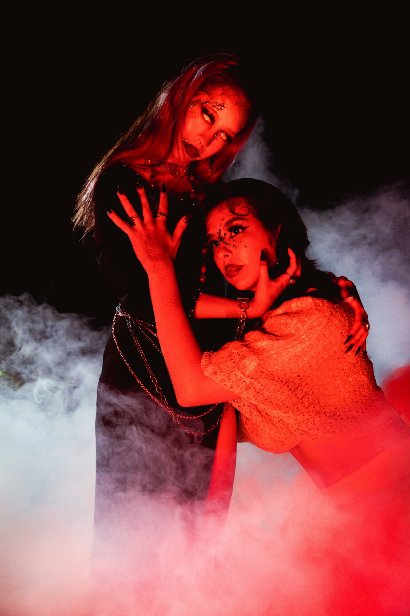 Two women pose with smoke and red lighting, one with hand on the other's face, in a dark setting.