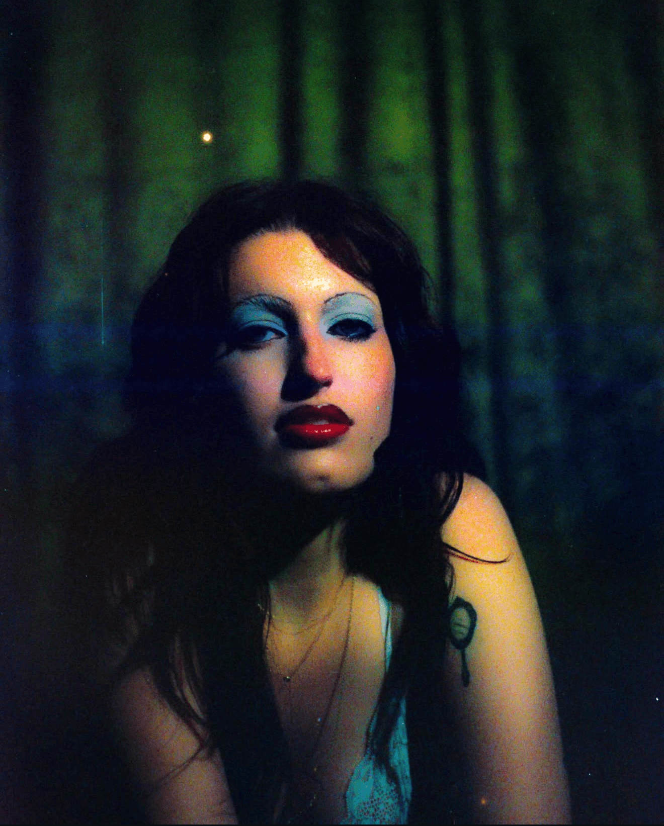 Portrait of a woman with dark hair and blue makeup, against a dark green background.