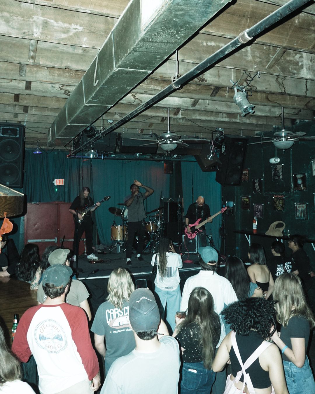 Band performs on stage with four musicians, audience watching in a dimly lit venue with exposed ceiling beams.