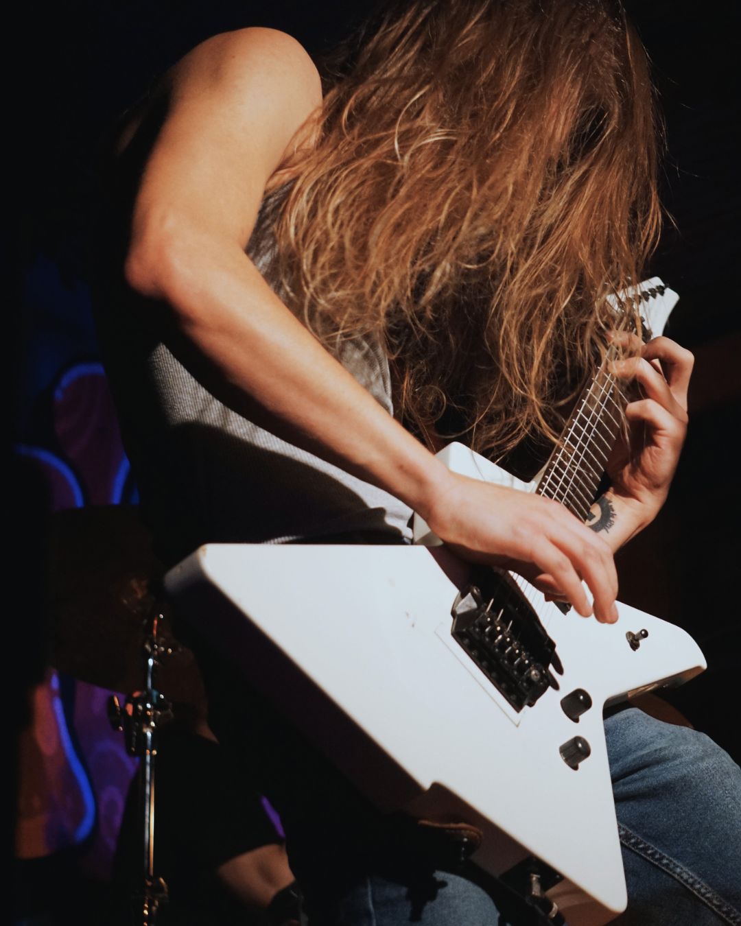 Person with long hair playing an electric guitar, head down, in a dark setting.