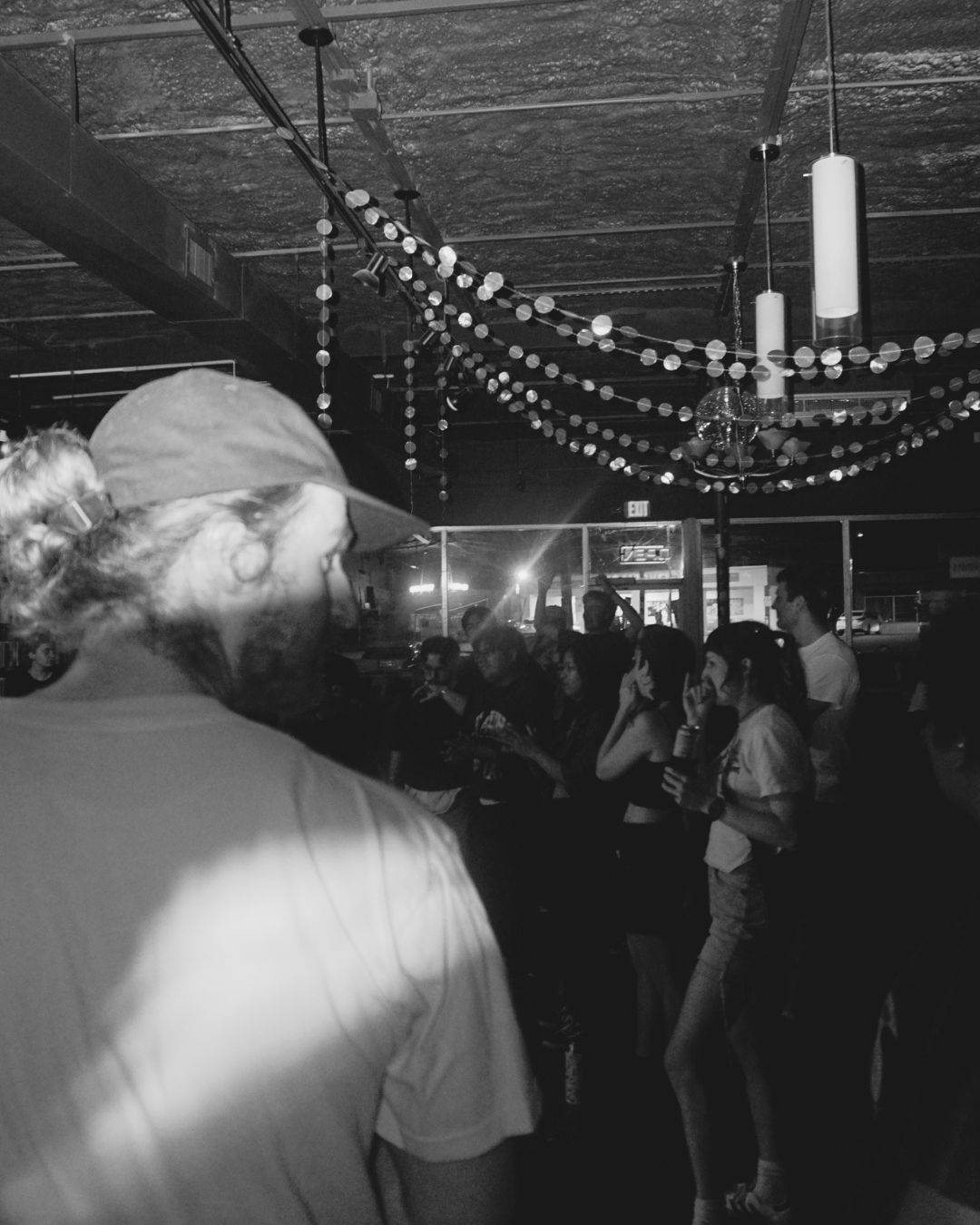People dancing and socializing in a decorated indoor space with hanging string lights and candles, black and white photo.