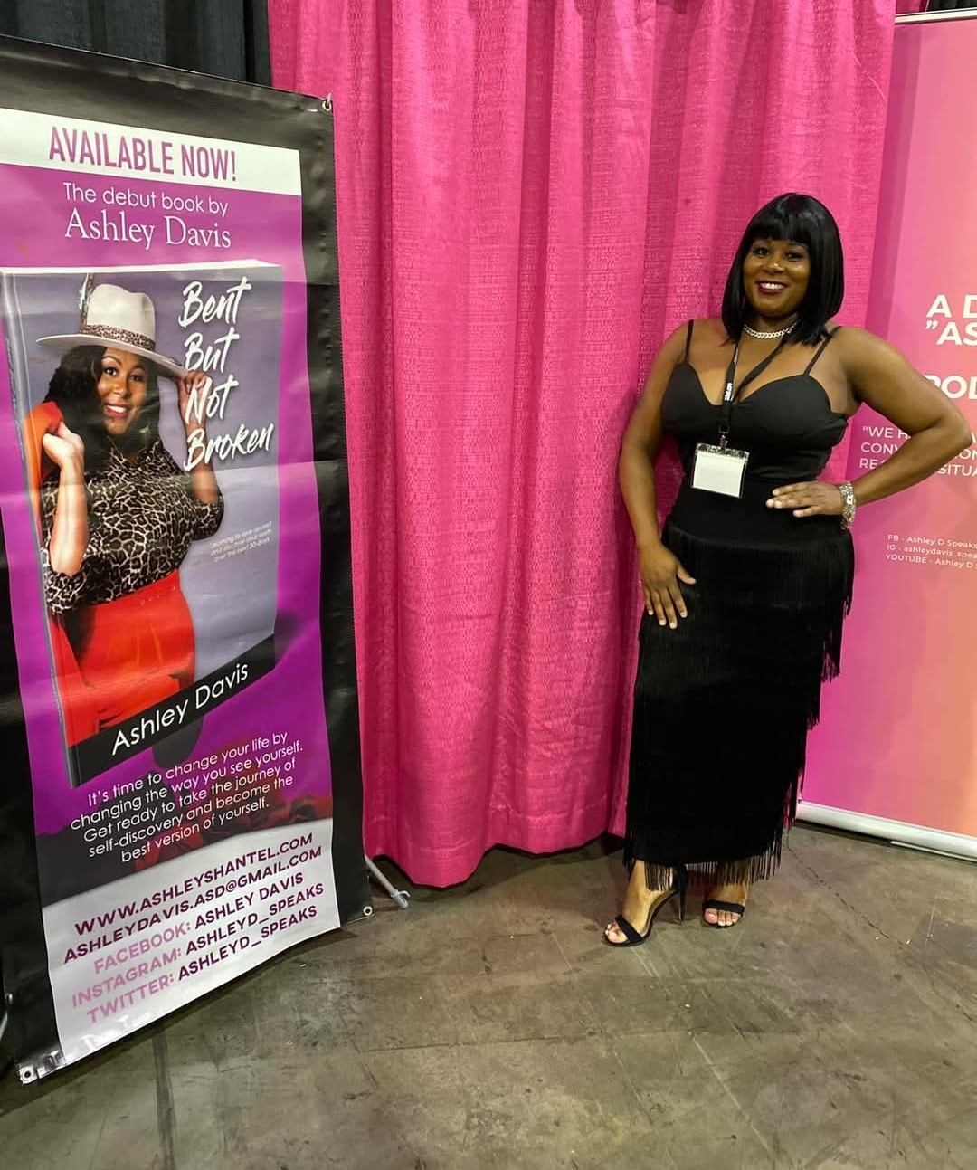 Woman with black hair and a black dress standing next to a pink curtain at a book event, smiling with hands on hips.