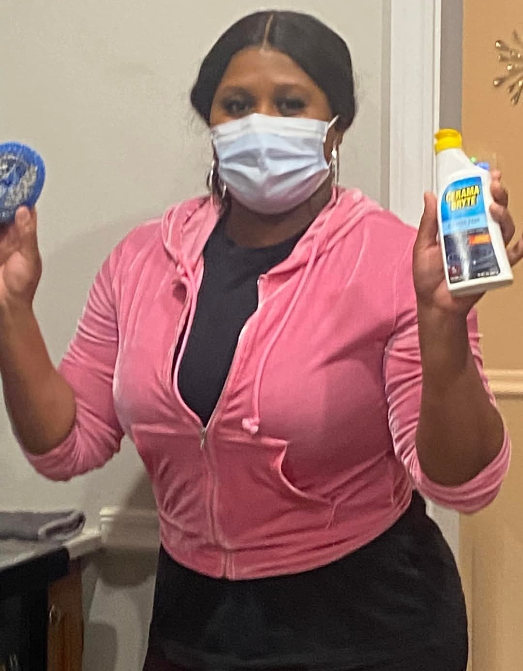 Woman wearing a face mask holding a bottle of cleaning product, standing indoors with a neutral background.