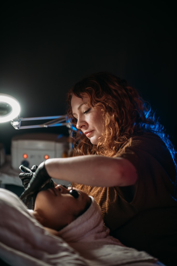 A woman with curly hair performs a cosmetic procedure on a person lying down, in a dimly lit room with a ring light.